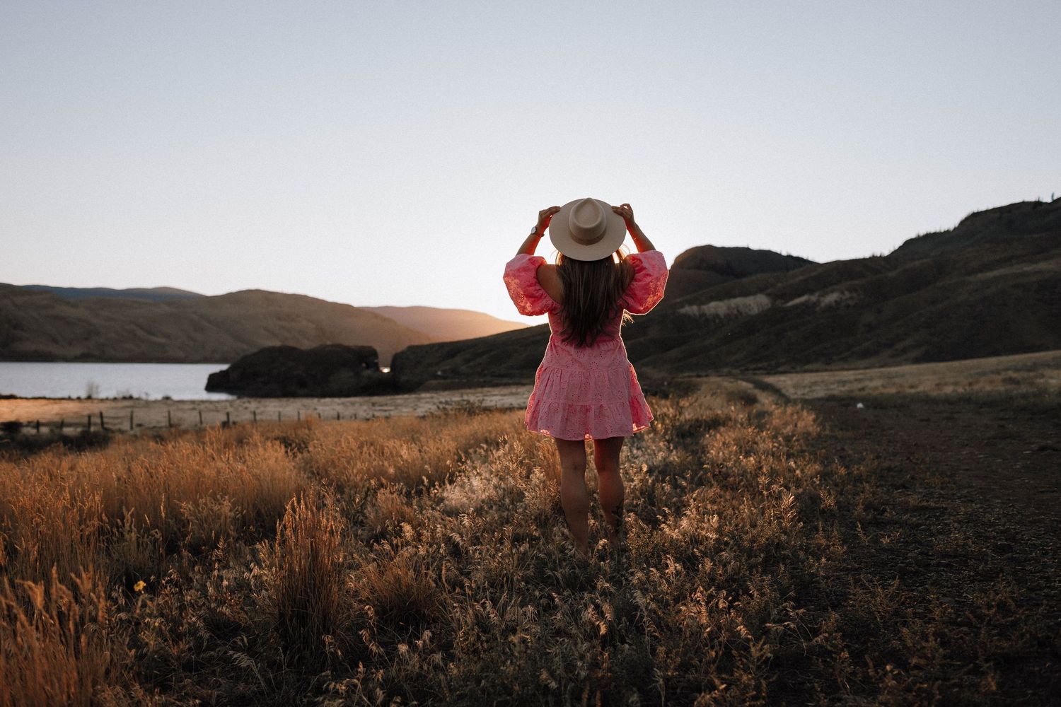 Person in a pink dress stands on hillside against sunset view of mountains and lake in golden hour.