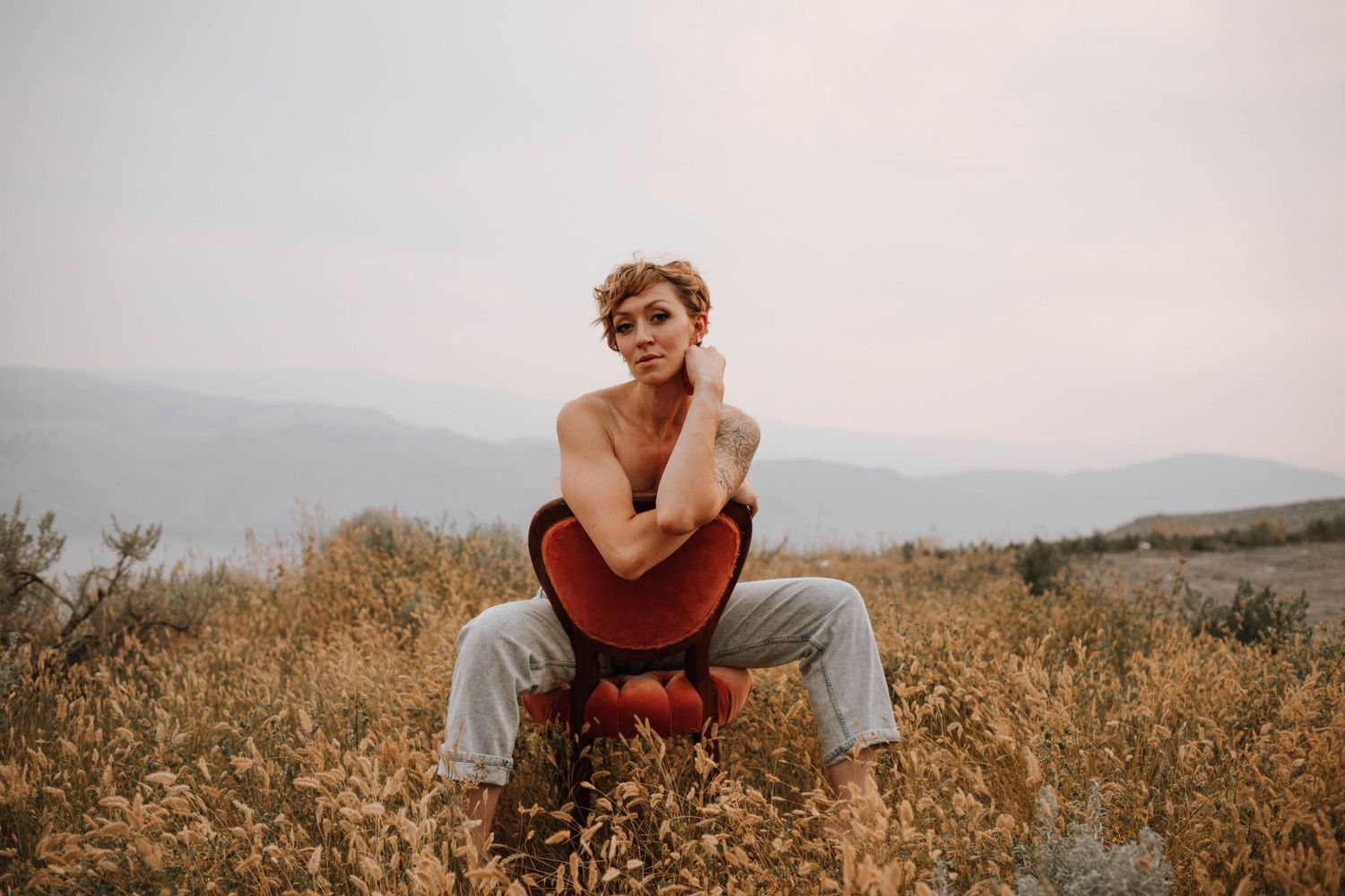 Person sitting in casual attire on dry grass against a misty mountain landscape at sunset.
