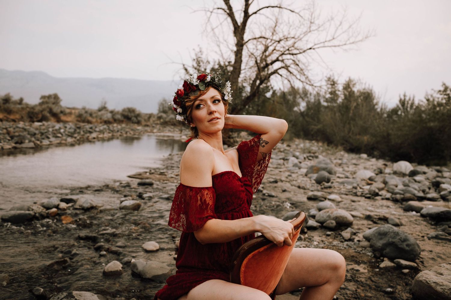Ethereal photoshoot featuring off-shoulder burgundy dress on rocky riverbed landscape.