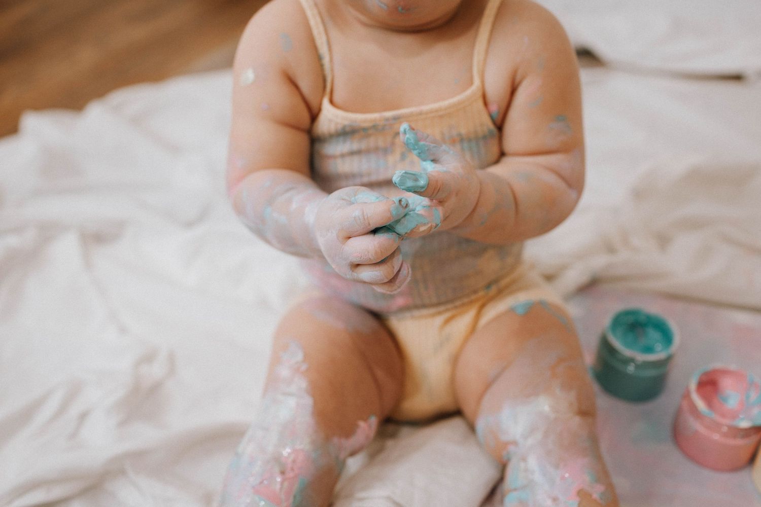 Baby in white onesie playing with colorful paints on a white sheet.