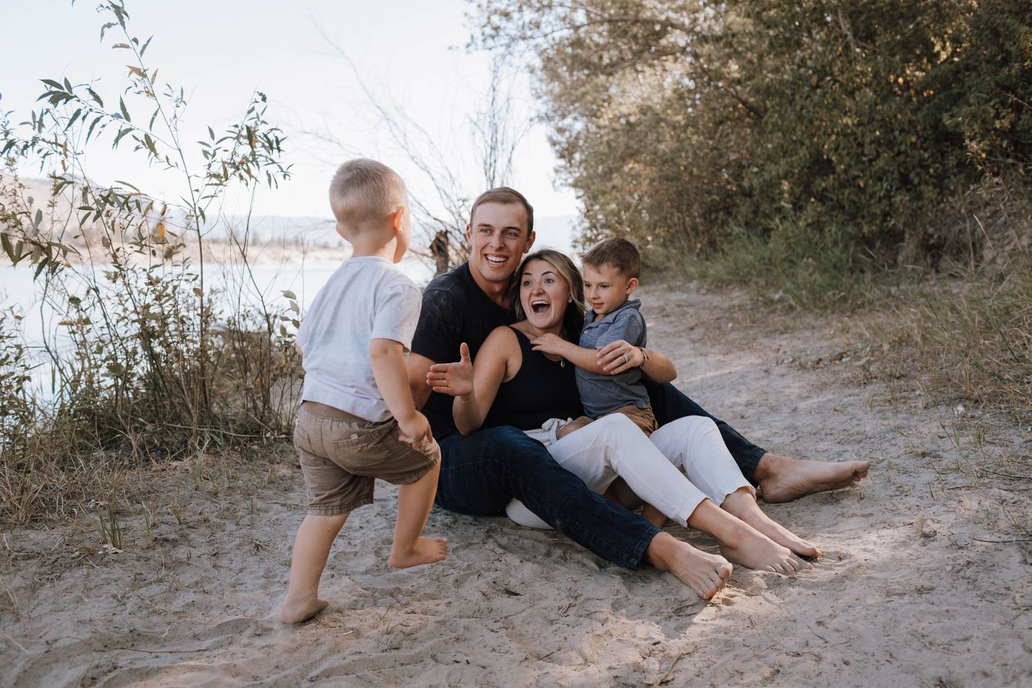 Family sharing candid laughter while lounging on a sandy trail near trees during a casual photo session.