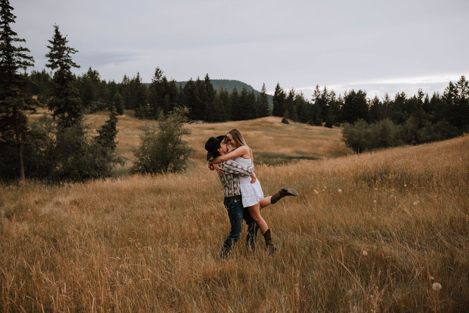 A joyful embrace captured in a rustic field with evergreen trees and mountains in the distance.