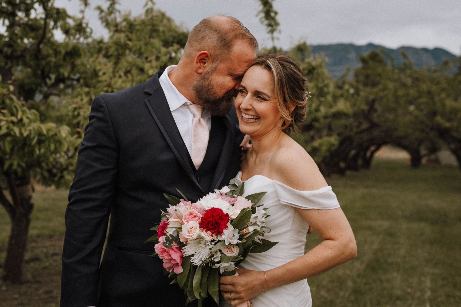 A newlywed couple sharing an intimate moment in an orchard while holding a pink and red floral bouquet.
