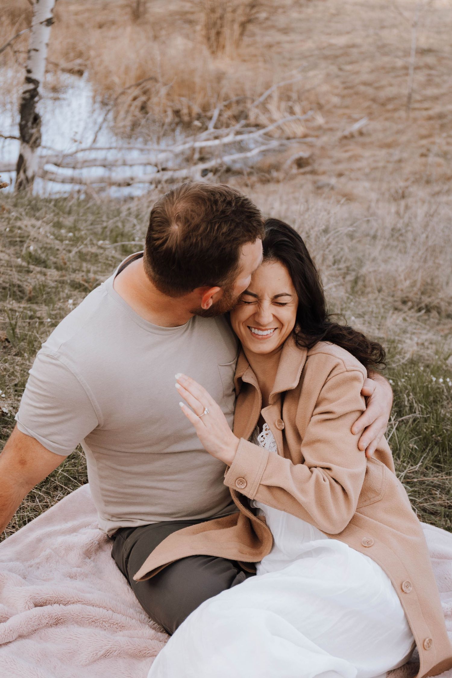 A couple shares an intimate moment while sitting on a pink blanket outdoors against a rustic backdrop.