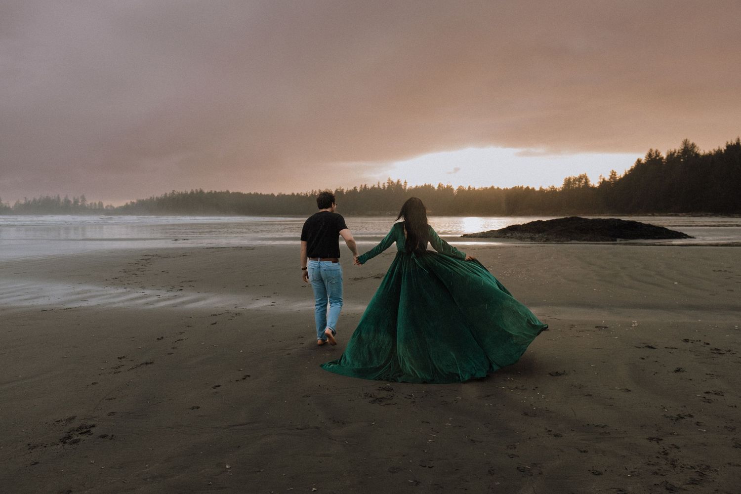 Couple holding hands on a misty beach at sunset, woman in flowing green gown, romantic coastal scenery.