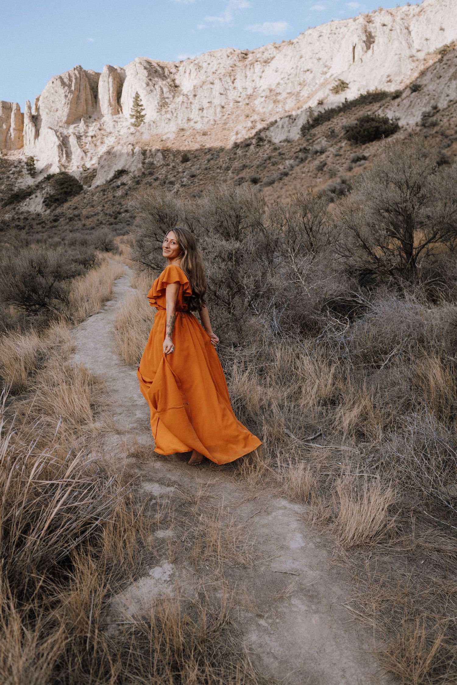 A person in a flowing orange dress walks along a winding desert trail beneath towering white cliffs at sunset.