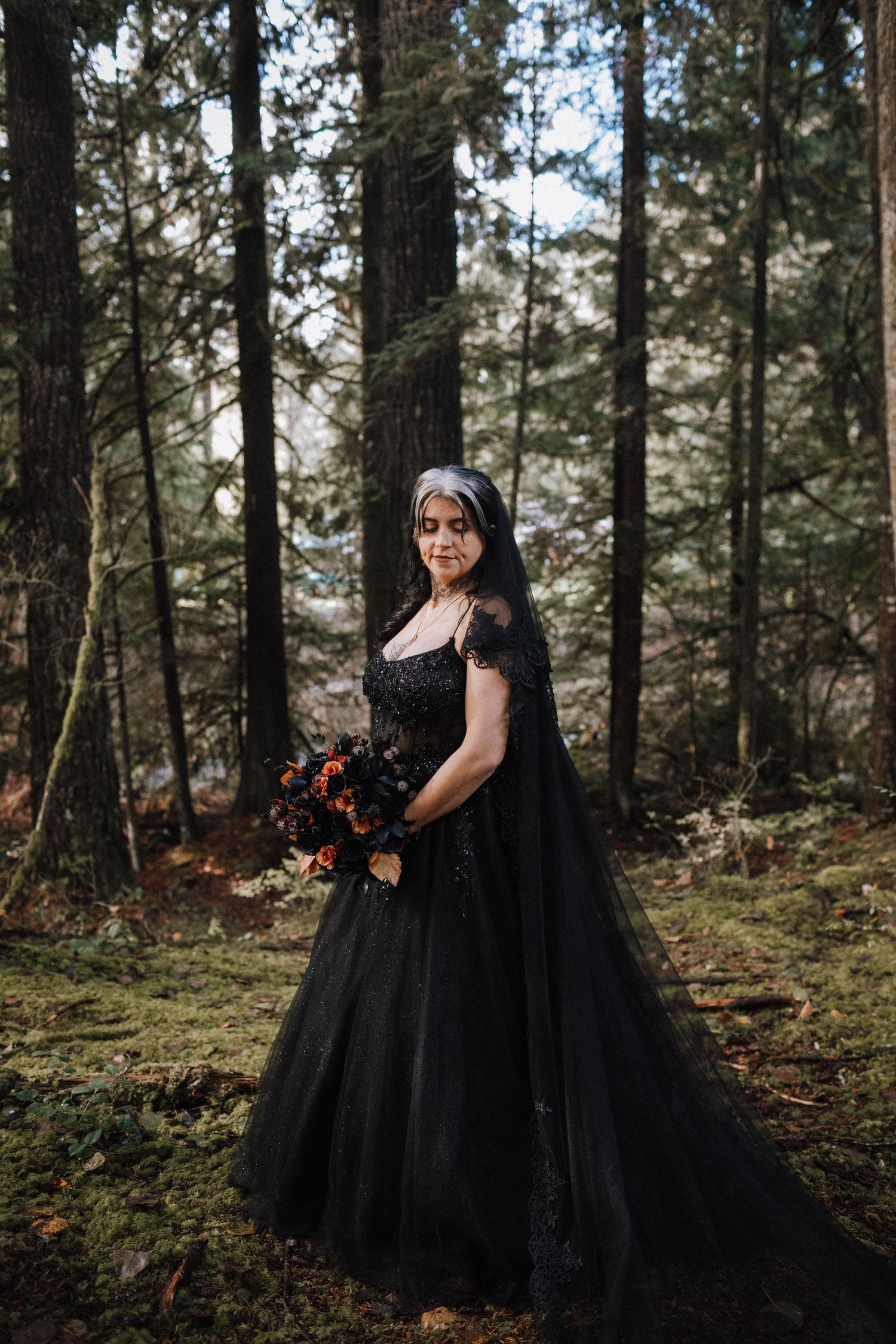 A person in a dramatic black ballgown poses in a mossy forest setting with orange florals.