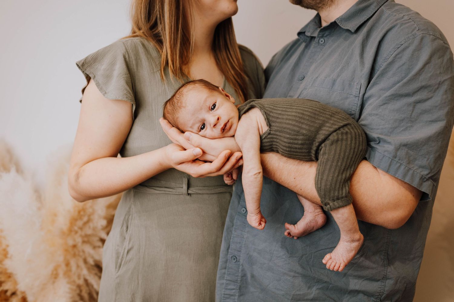 A sleeping baby in knit pants is cradled by parents wearing neutral-colored clothing against a light background.