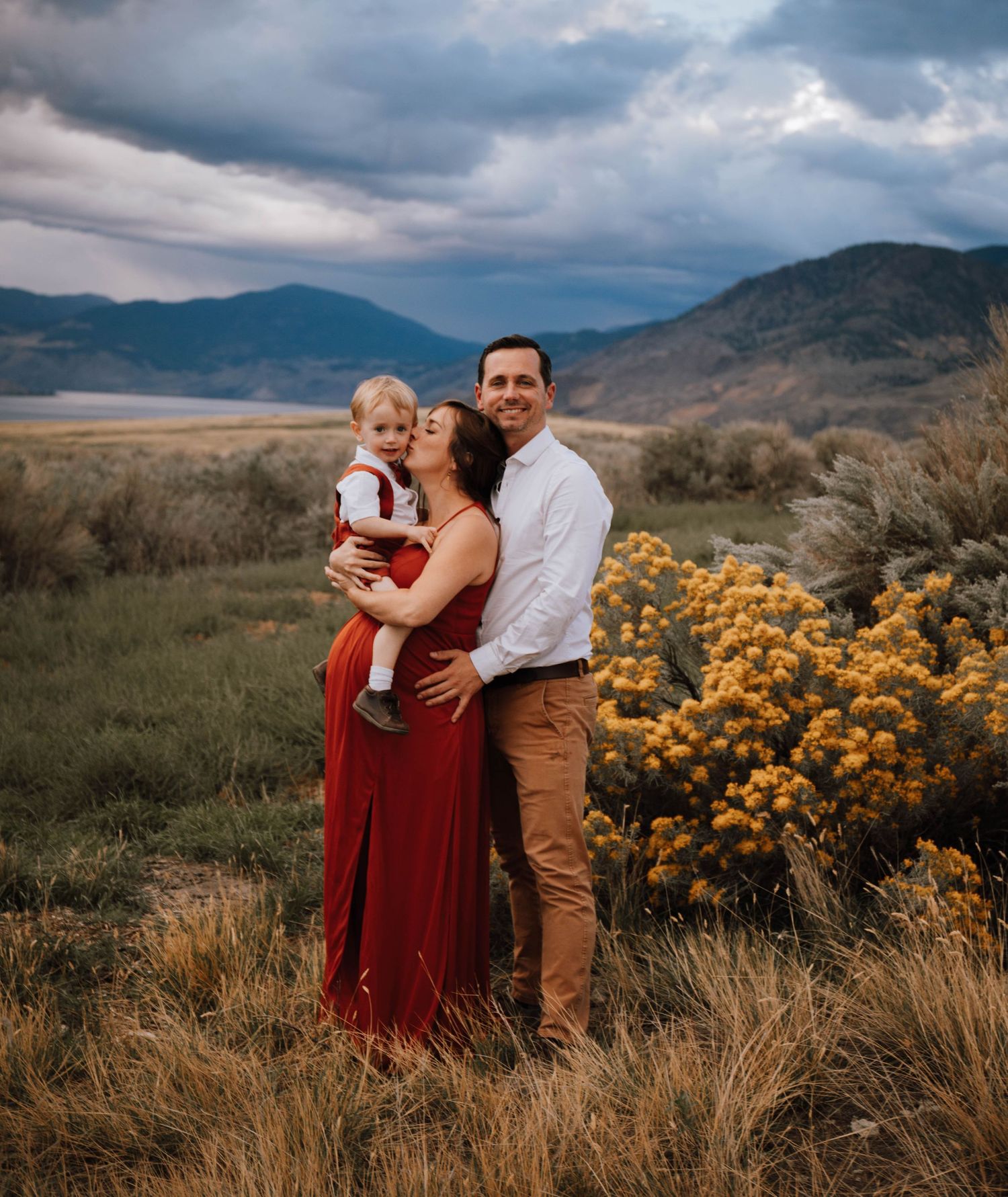 A family poses together on a hillside with dramatic cloudy skies and yellow wildflowers in the background.