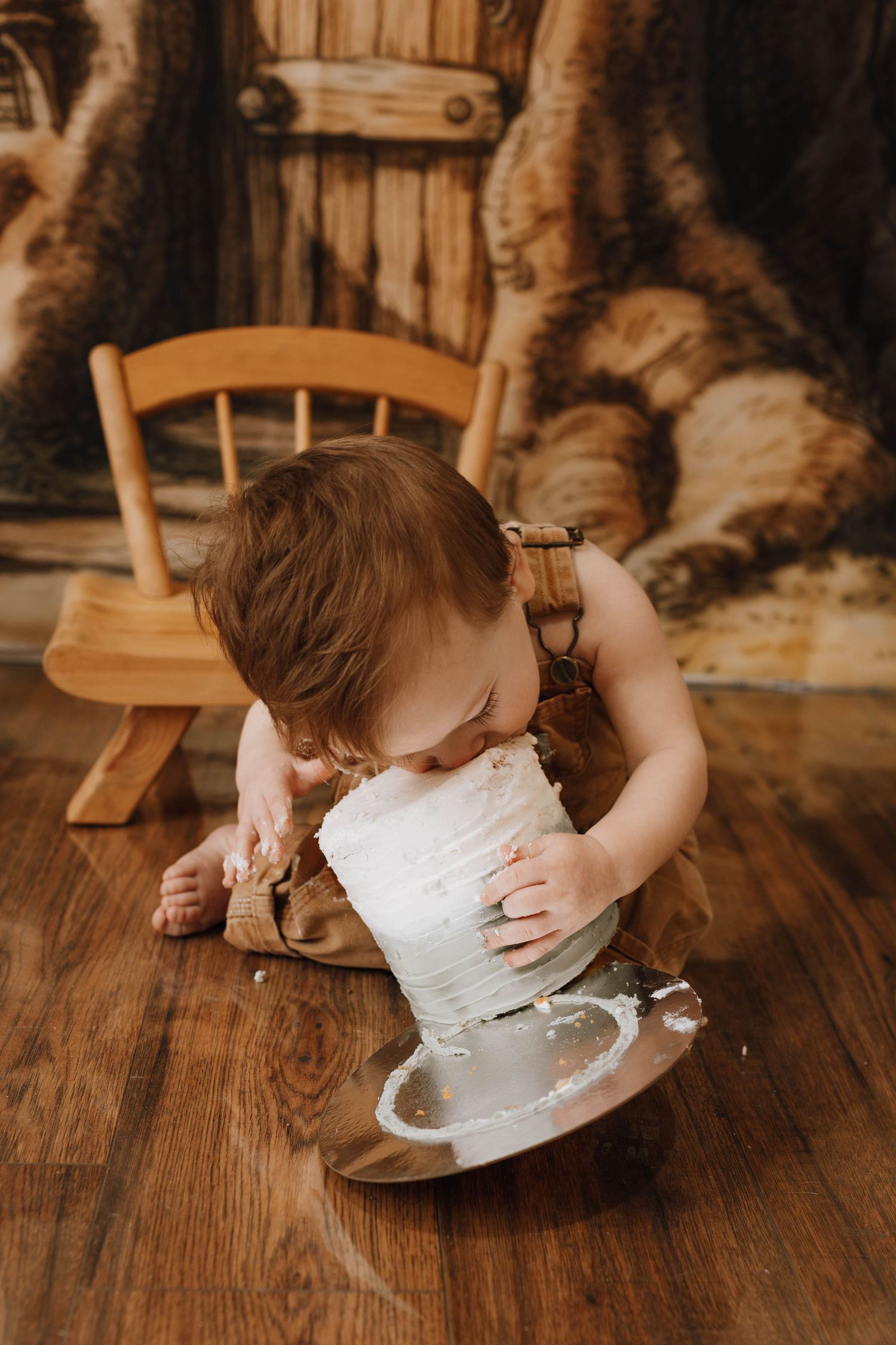 Baby in overalls playing with white cake on wooden floor against rustic backdrop.