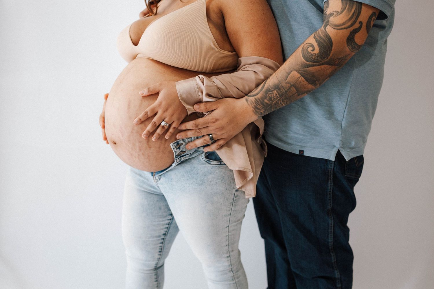 Tattooed arms embrace a pregnant belly while wearing casual jeans and neutral colors against a white wall.