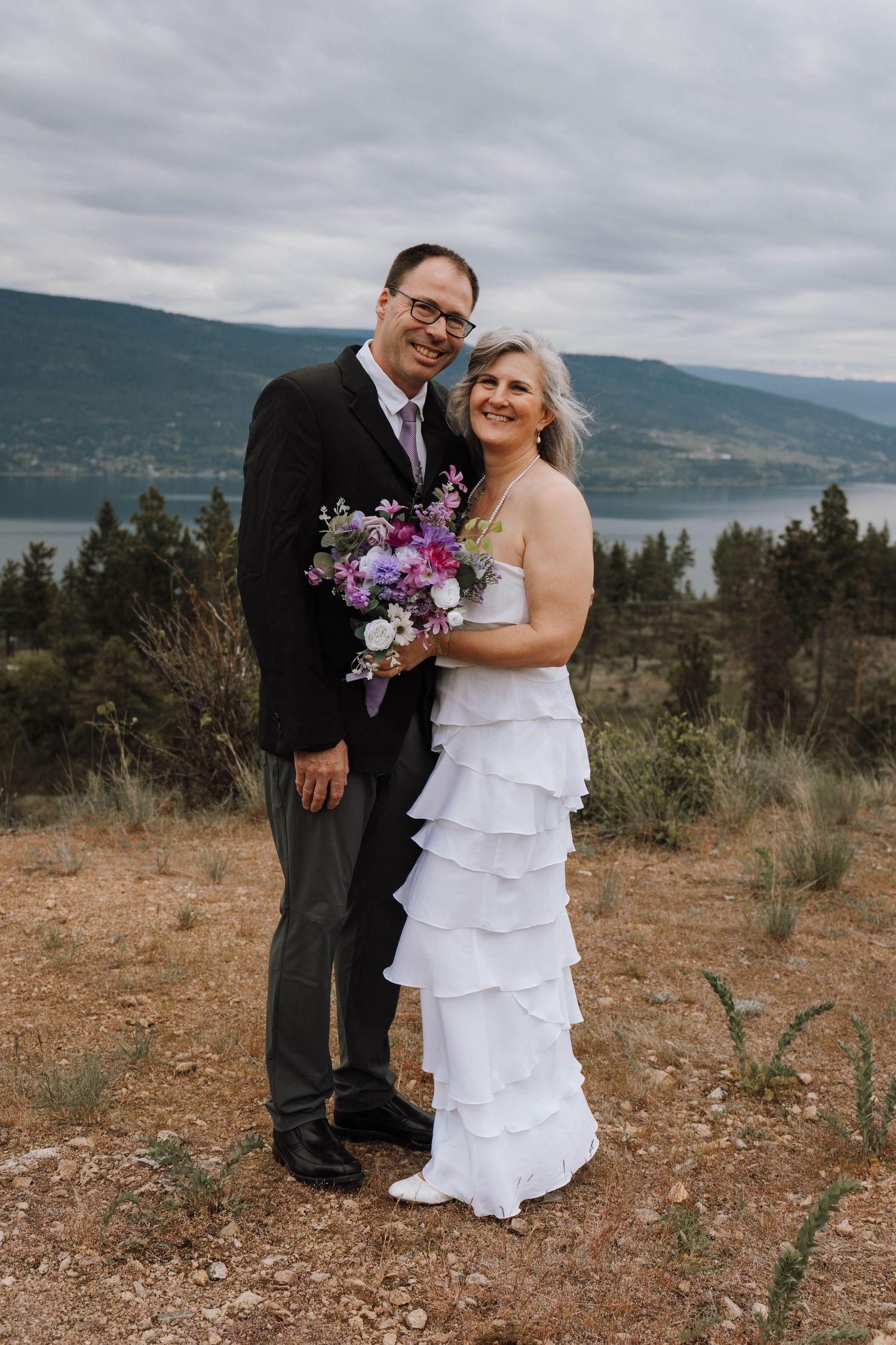 A couple stands overlooking a lake and mountains on their wedding day, with the bride holding a purple and pink bouquet.