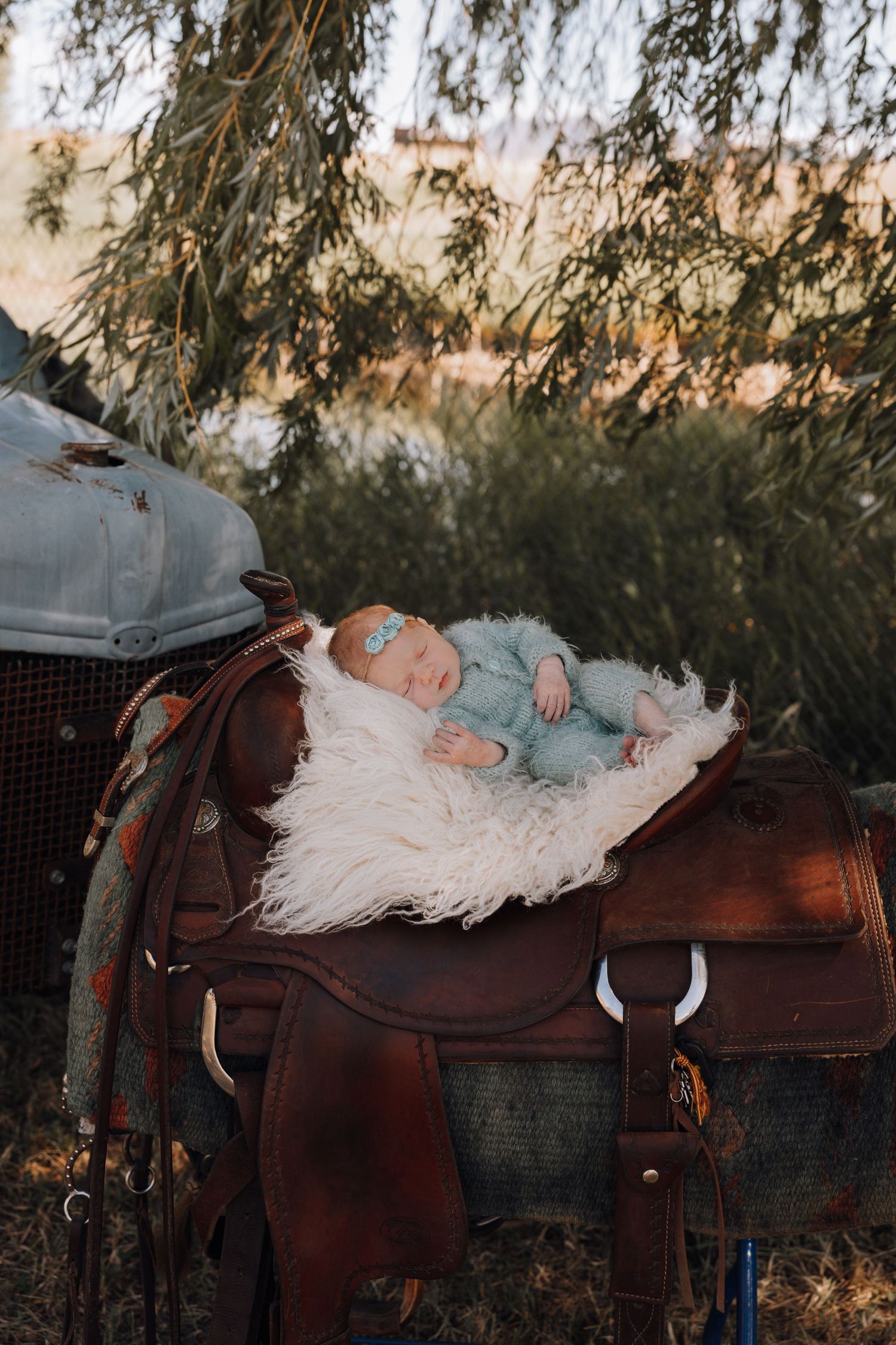 Sleeping newborn baby in blue outfit resting on fluffy white fur atop a brown leather western saddle outdoors.