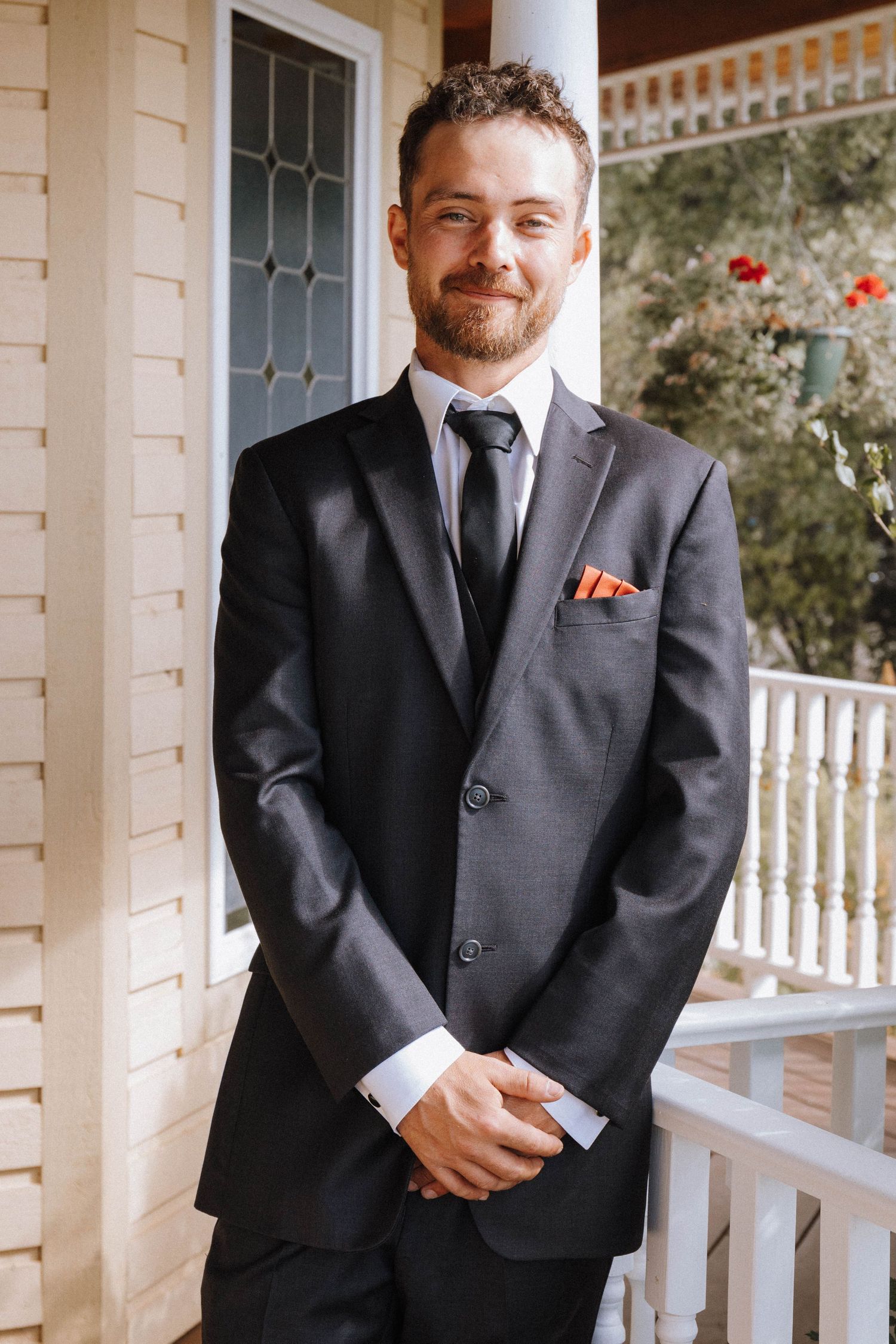 Groomsman in a dark suit with a red boutonniere smiles while standing on a white porch with columns.