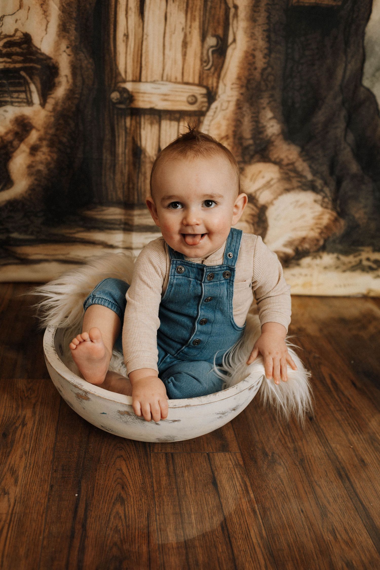 Smiling baby wearing overalls seated in white bowl against painted woodland scene.
