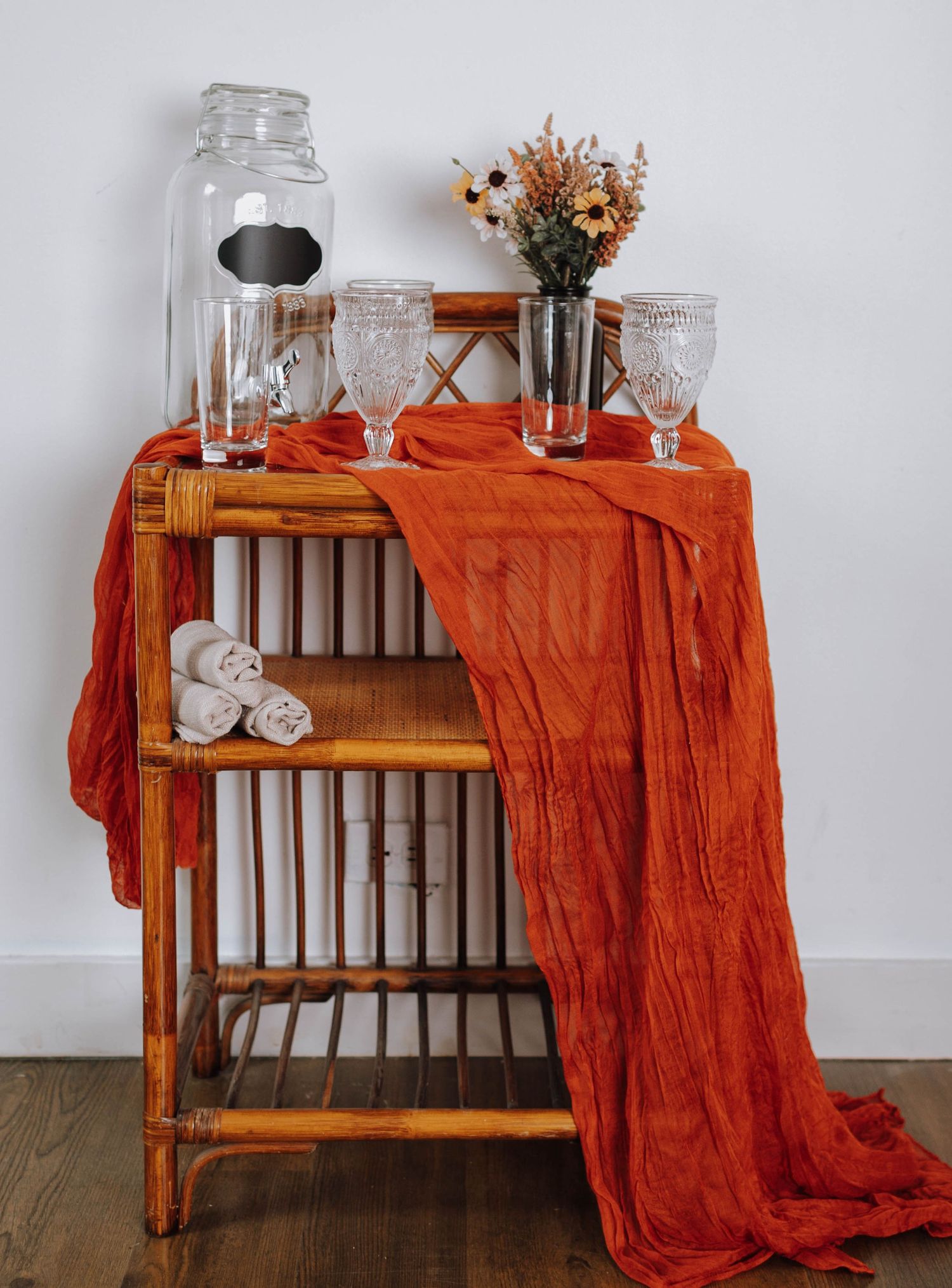 Wooden shelf decorated with glassware, orange fabric, and dried flowers against white wall.