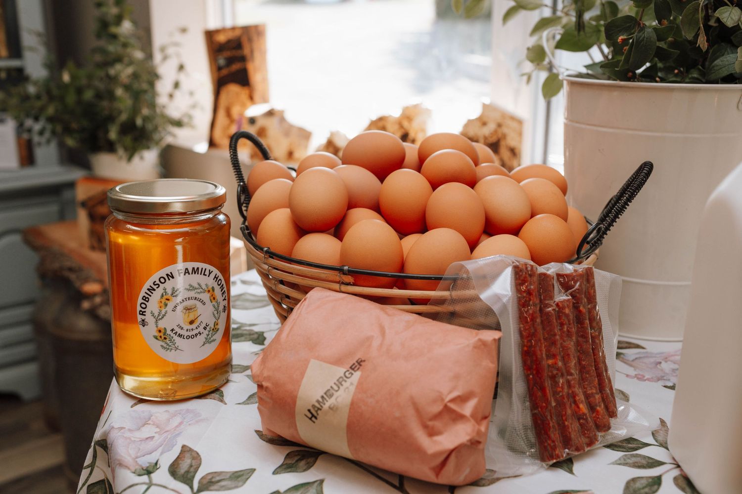 Fresh eggs, honey jar, and wrapped goods arranged on patterned tablecloth in rustic kitchen setting.