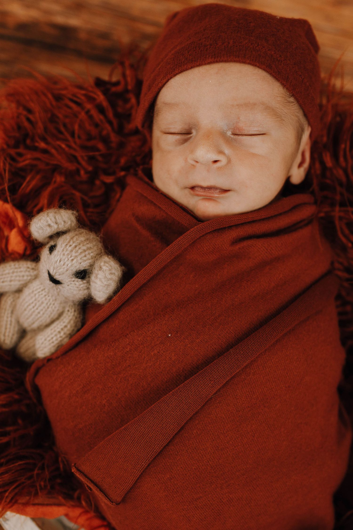 Sleeping baby wrapped in burgundy swaddle with knit stuffed animal on red textured blanket.
