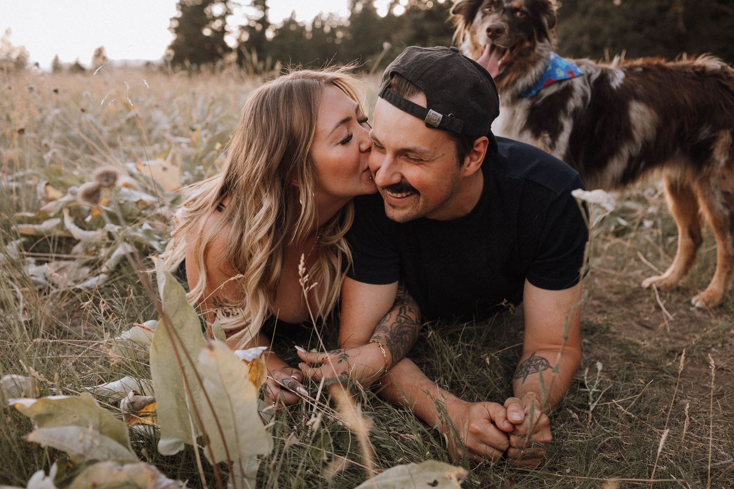 A couple shares an intimate moment lying in a field of tall grass with their dog visible in the background.