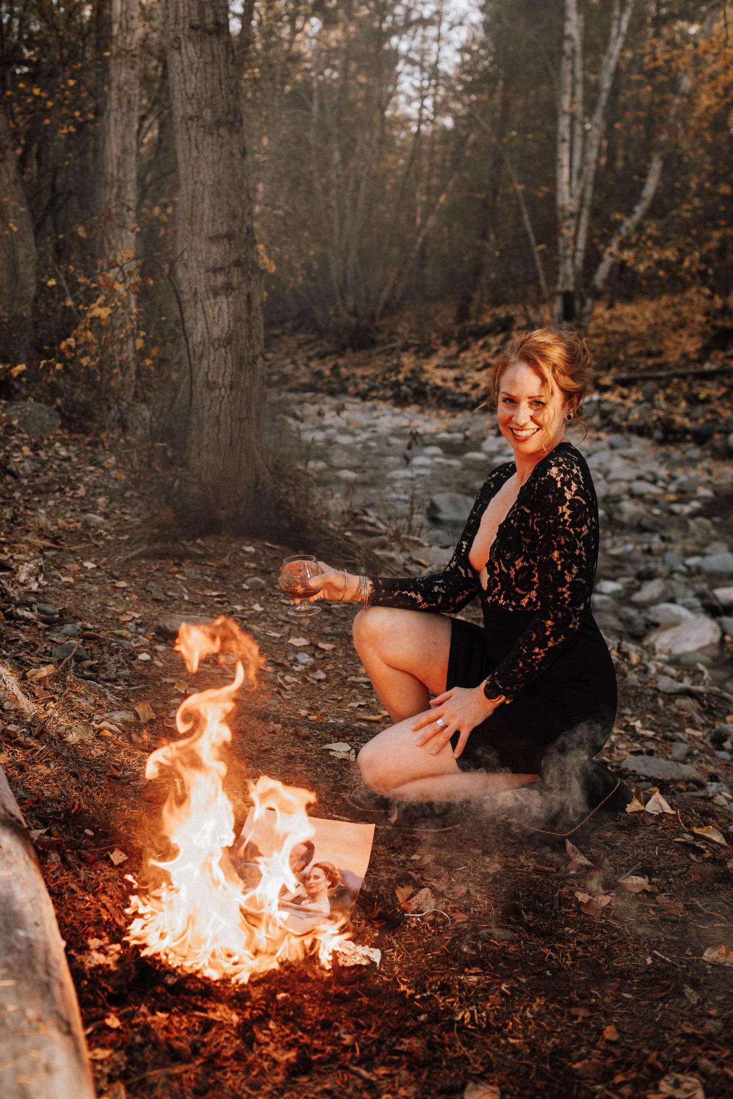 A person in a black lace dress sits by a warm campfire in an autumn forest setting with rocks and fallen leaves.