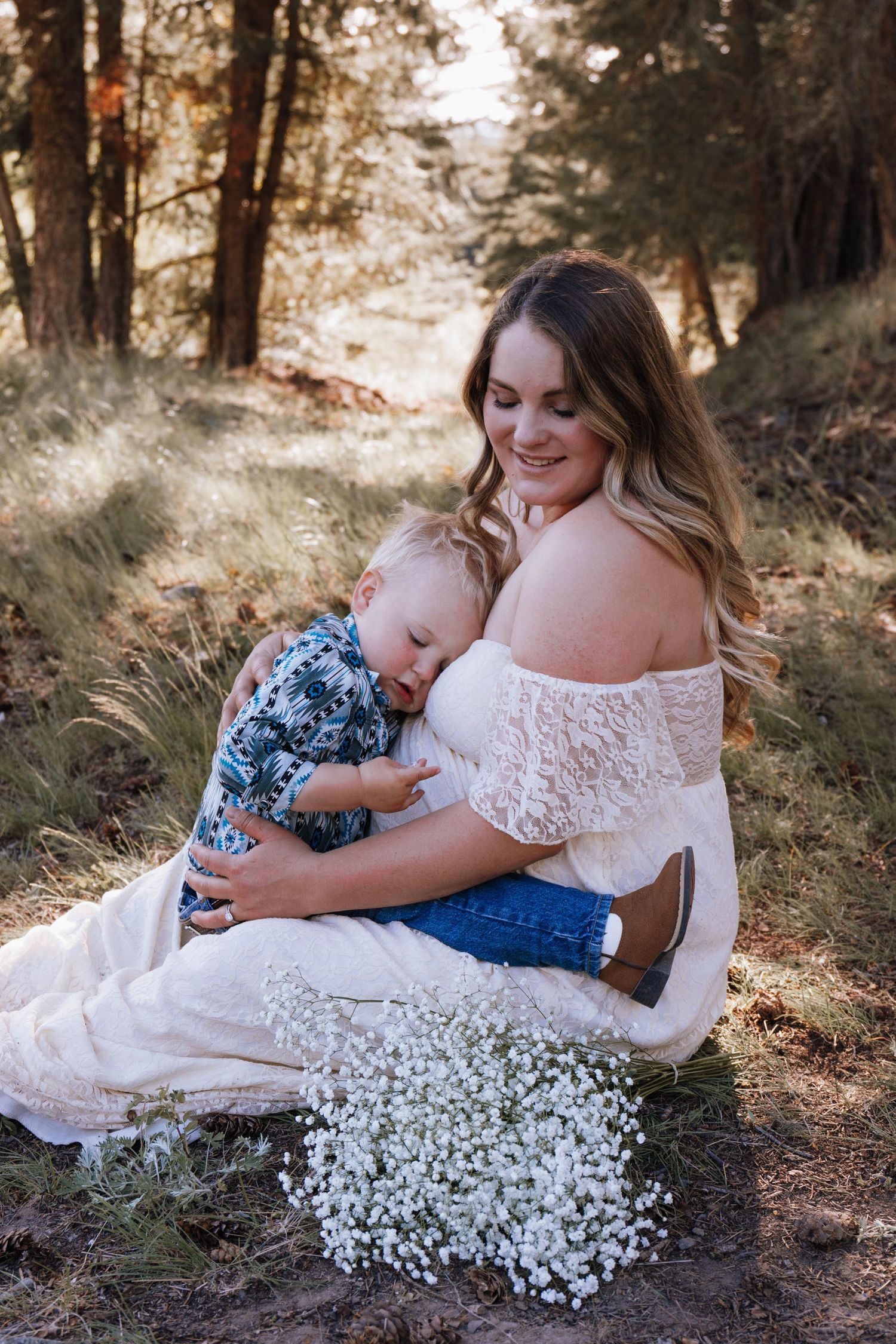 Parent and baby share tender moment in sunlit forest setting with white lace dress and baby's breath flowers.
