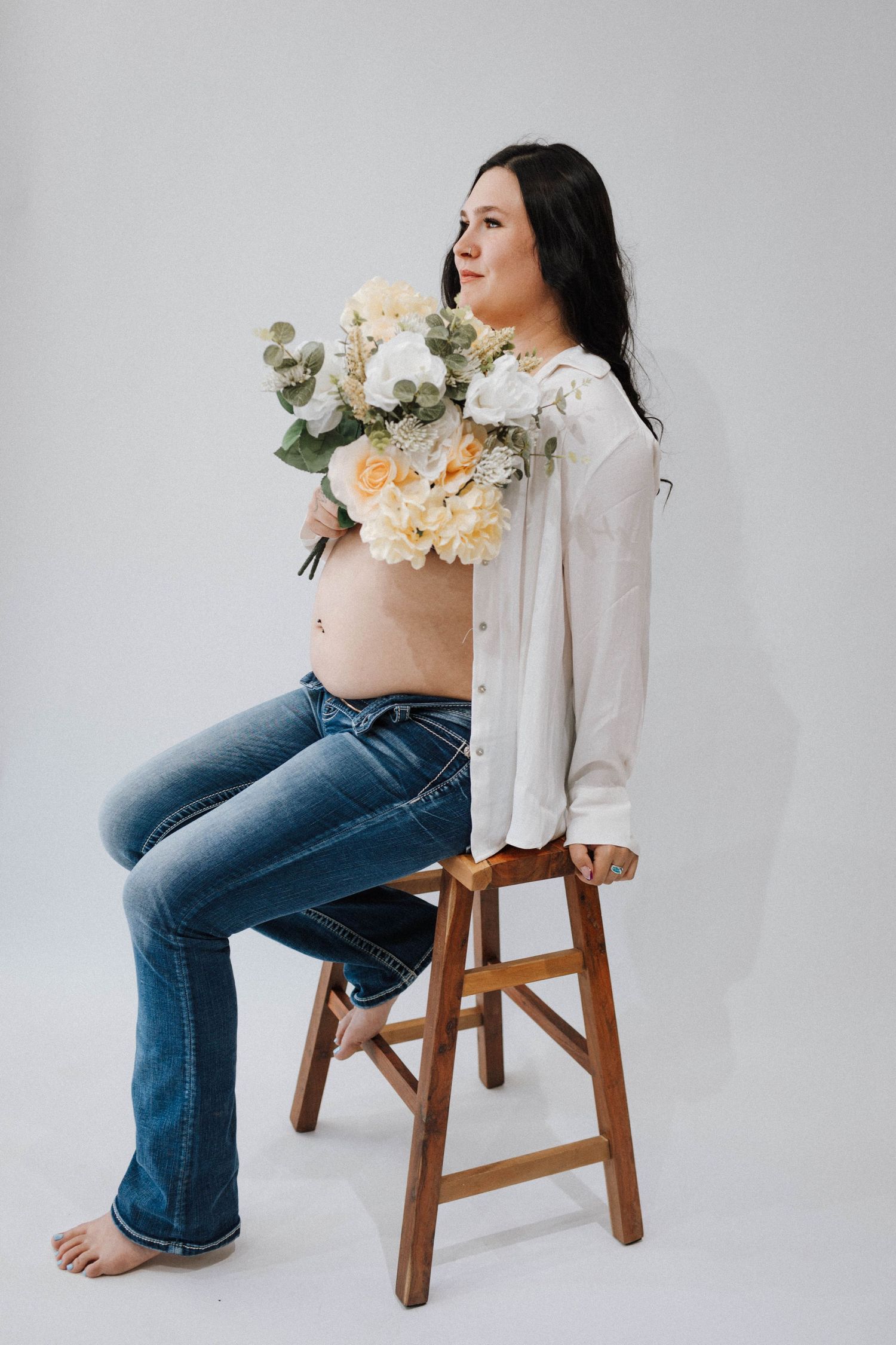 Minimalist maternity photo shows expectant mother in white blouse and jeans sitting on wooden stool holding floral bouquet.