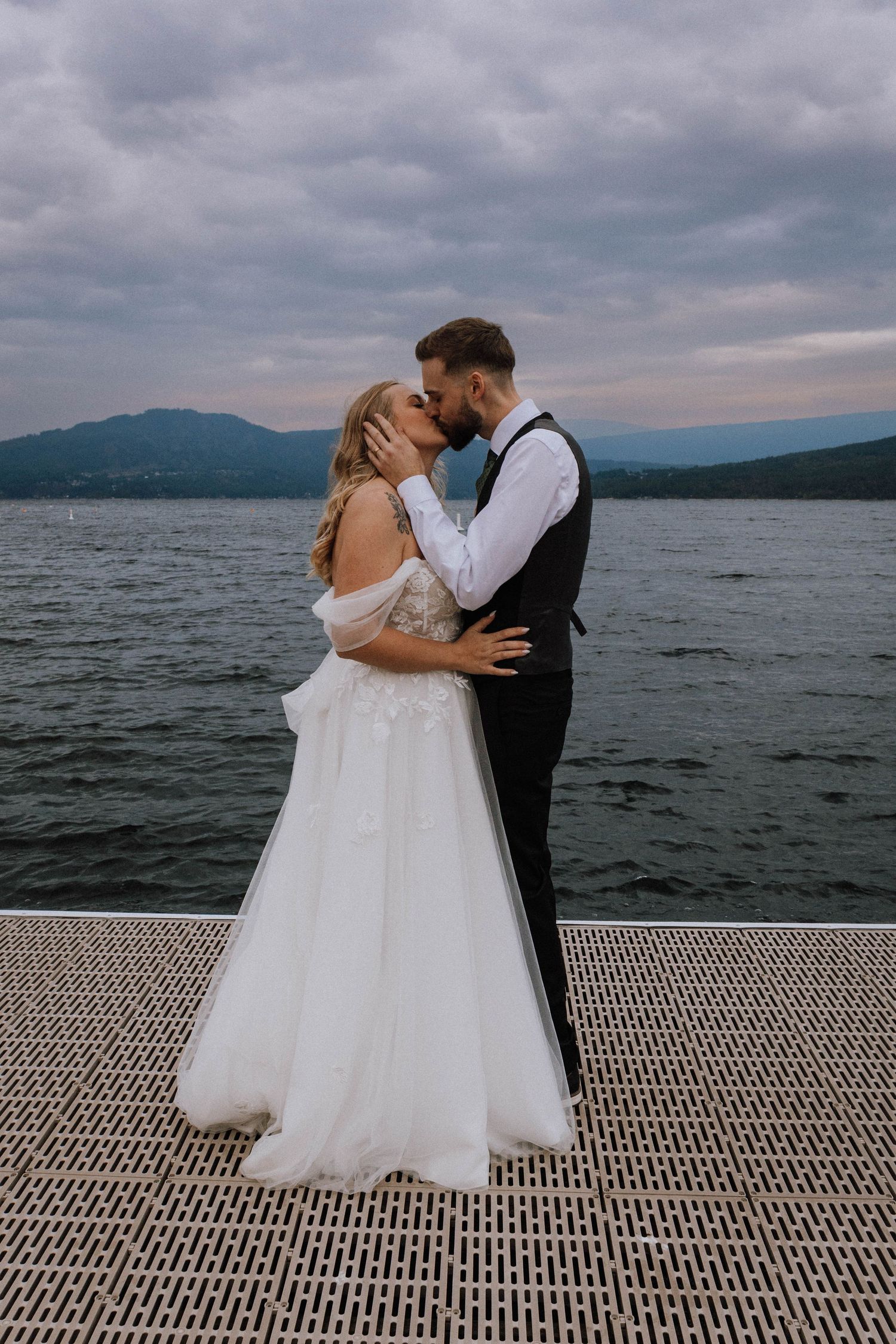 A couple shares a romantic kiss on a dock overlooking a misty lake with mountains in the background.