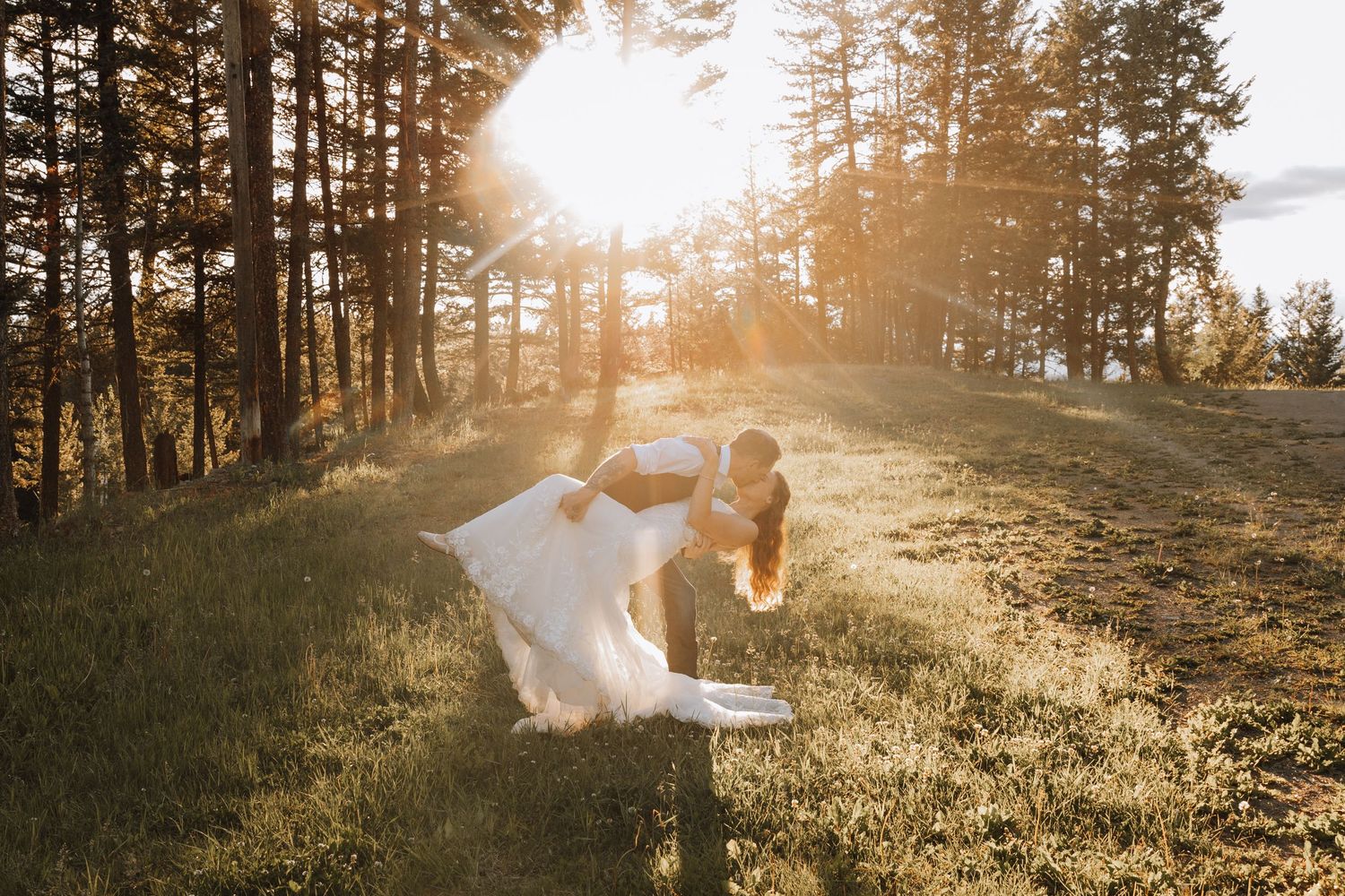 Couple sharing a romantic dip kiss in a sunlit forest meadow during golden hour wedding photography.