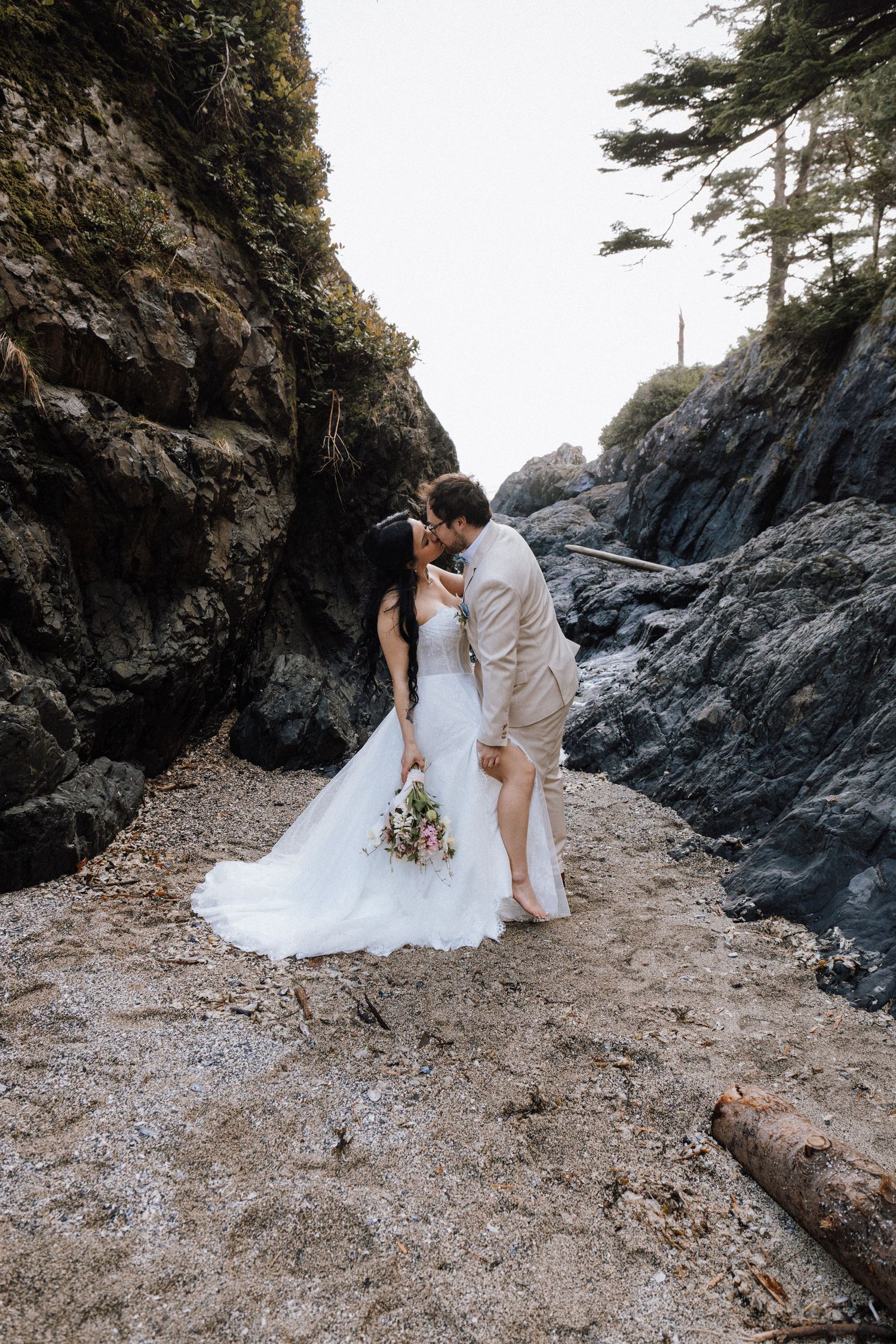 Bride and groom kissing on a rocky beach cove, bride holding a bouquet in a white gown, groom in a tan suit.