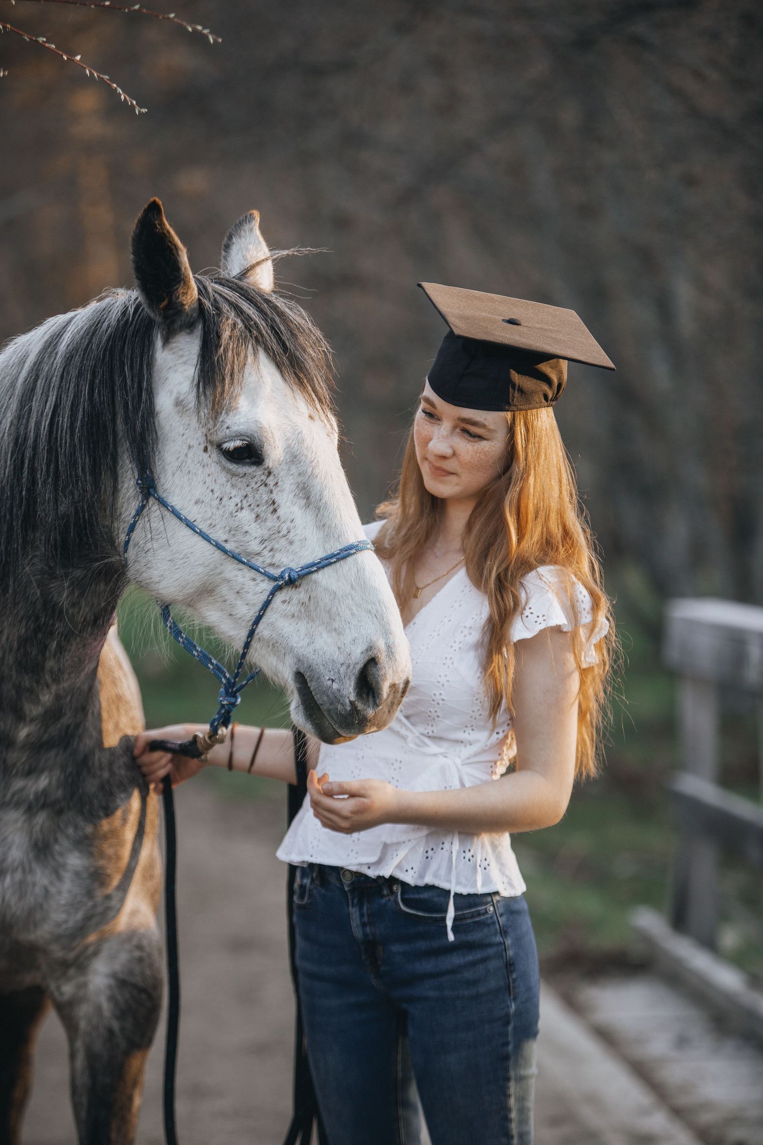 Graduate in white top and graduation cap stands with gray horse outdoors.