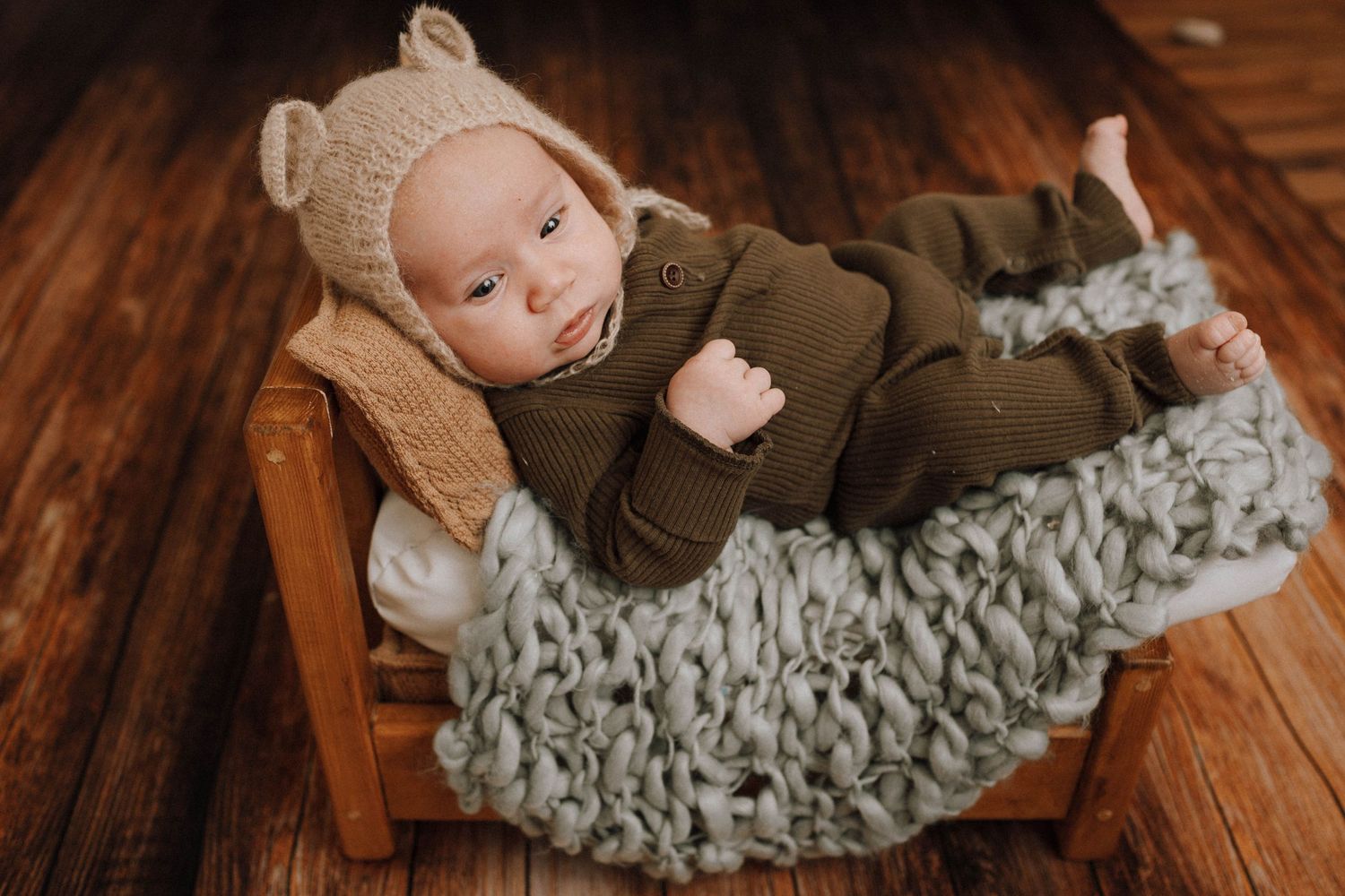 Sleeping baby wearing bear hat bonnet rests on textured blanket in wooden bed.