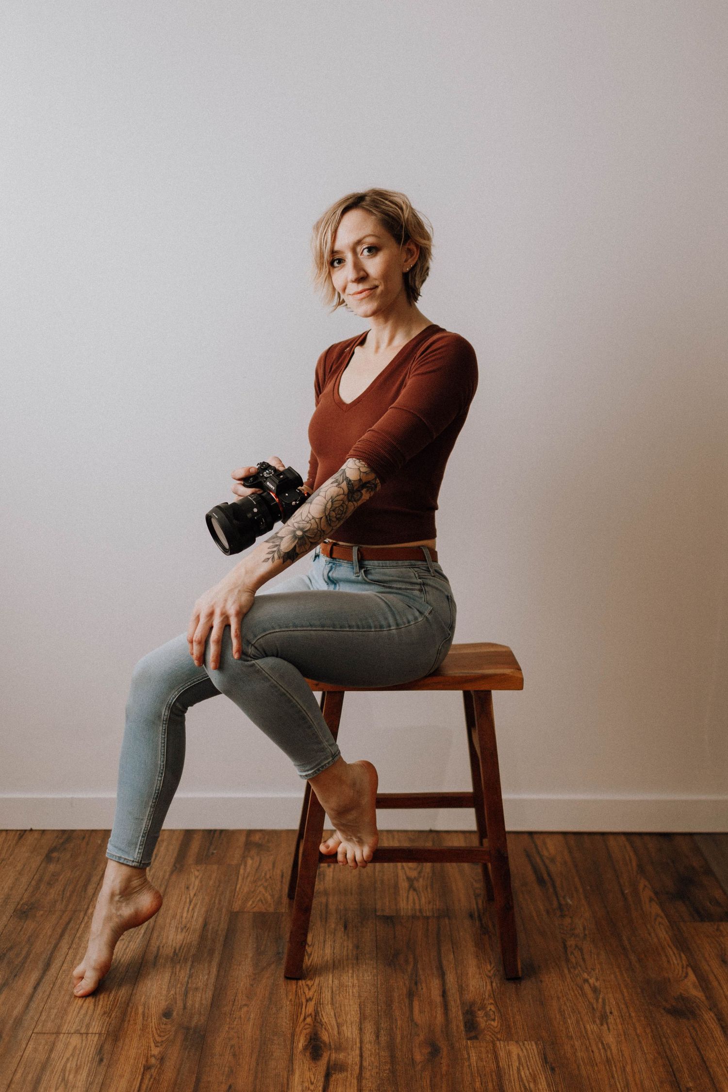 Professional photographer in brown sweater and jeans sits casually on wooden stool while holding digital camera.