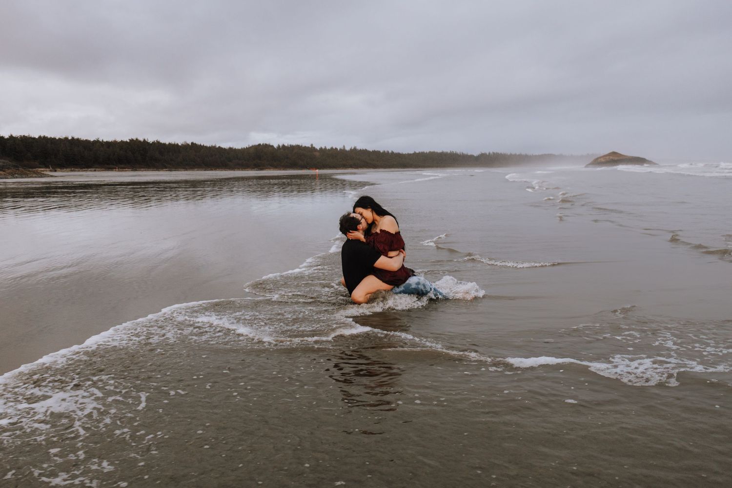 Couple embracing in ocean waves on a cloudy beach, surrounded by misty shoreline and forested hills.