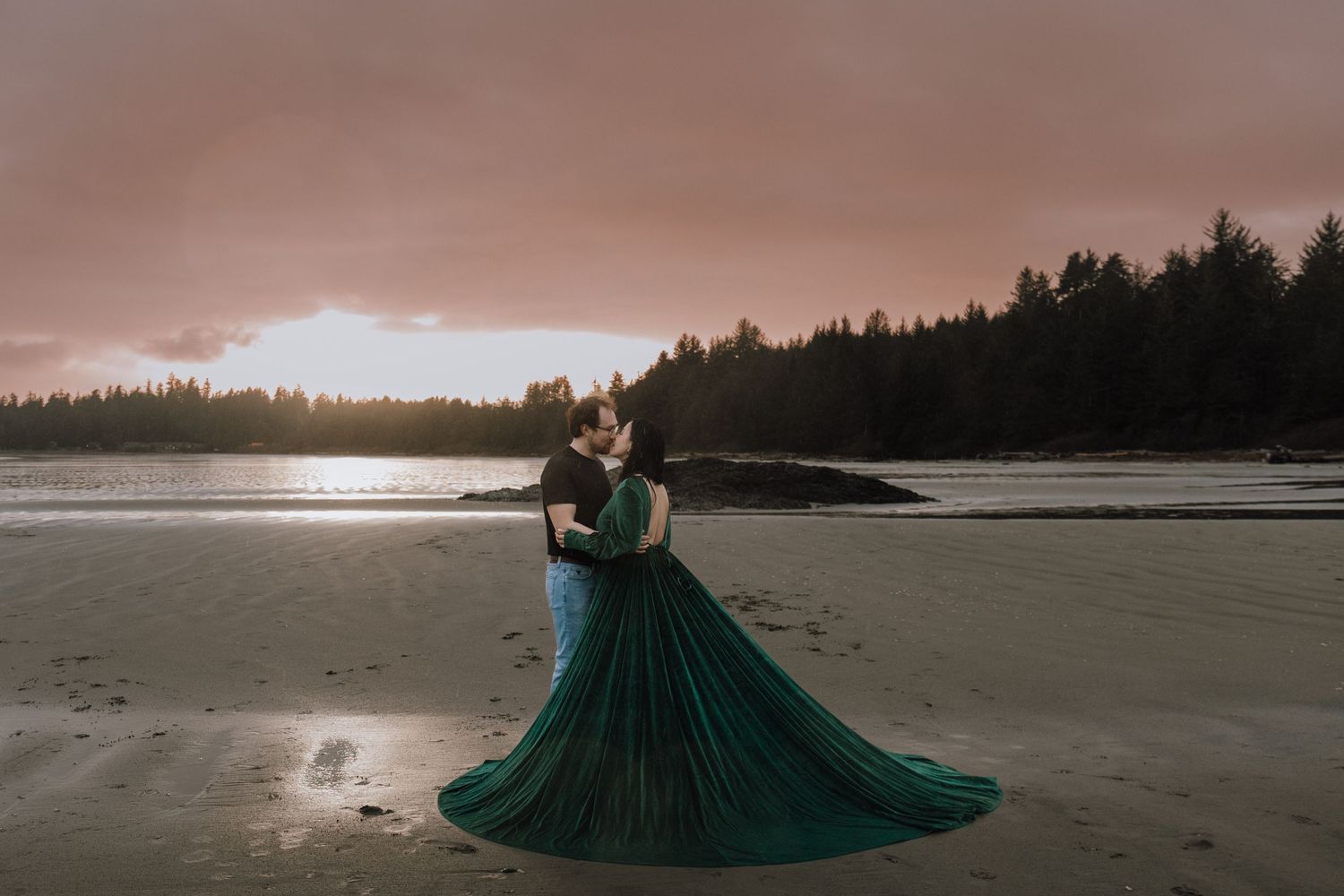Couple embracing on a beach at sunset, woman in flowing emerald green gown, dramatic sky and forest backdrop.