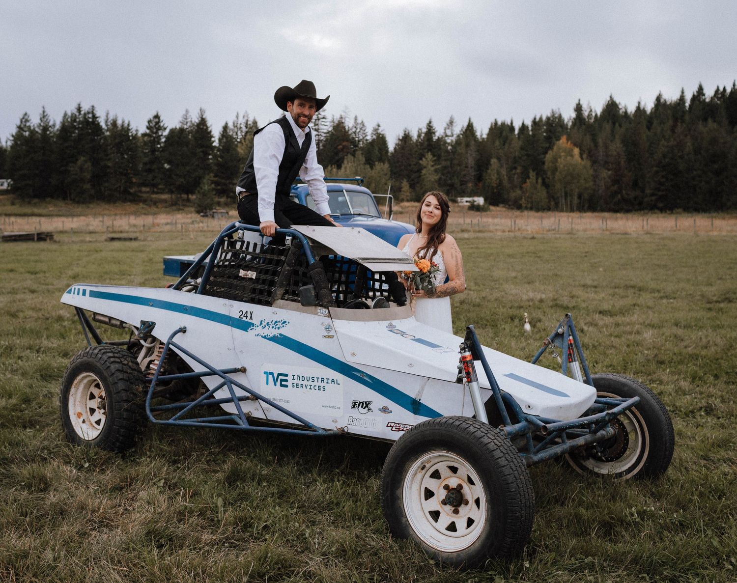 A couple poses together on a white off-road racing vehicle in a field with evergreen trees in the background.
