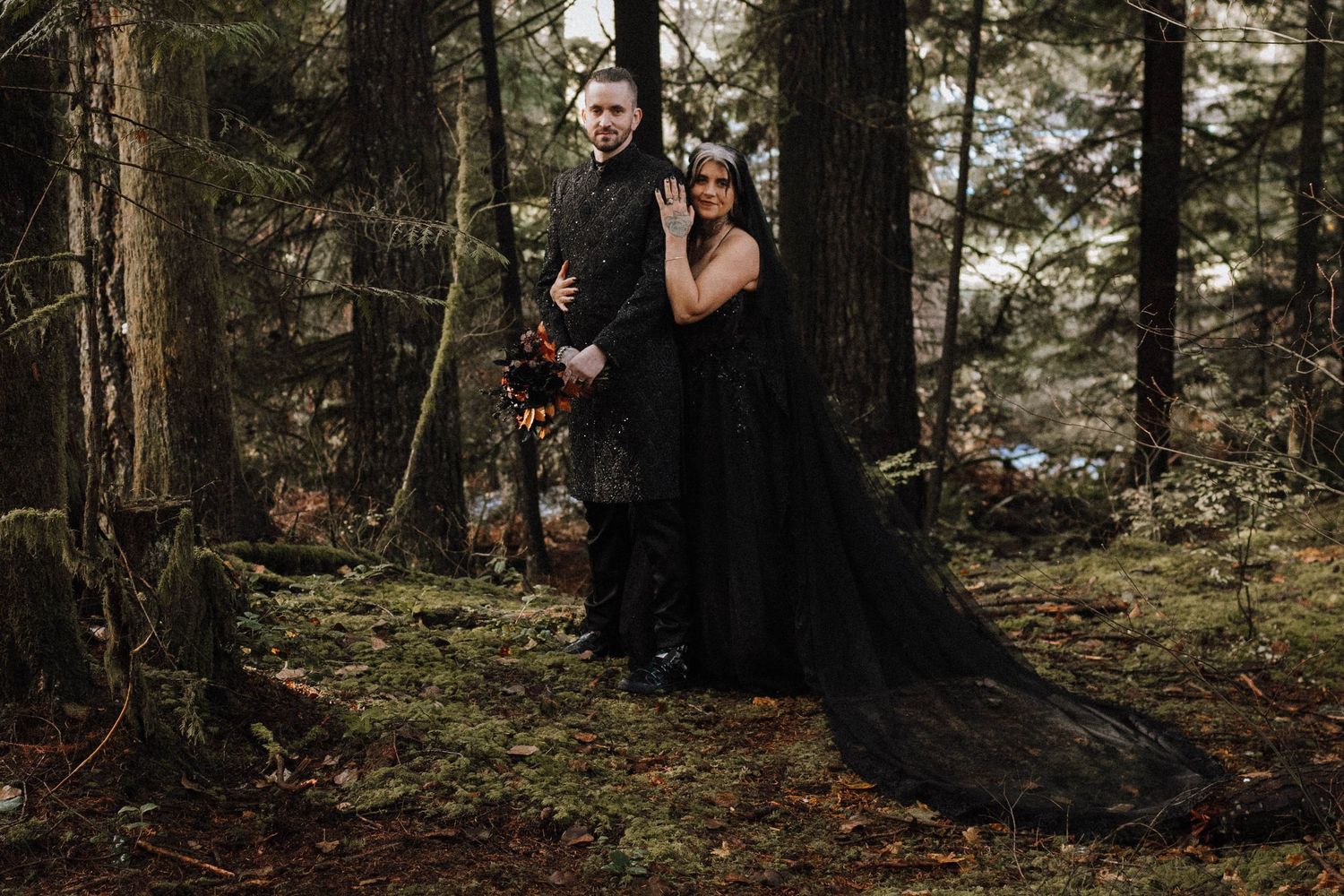 Gothic wedding couple in dramatic black attire pose in a moody forest setting.