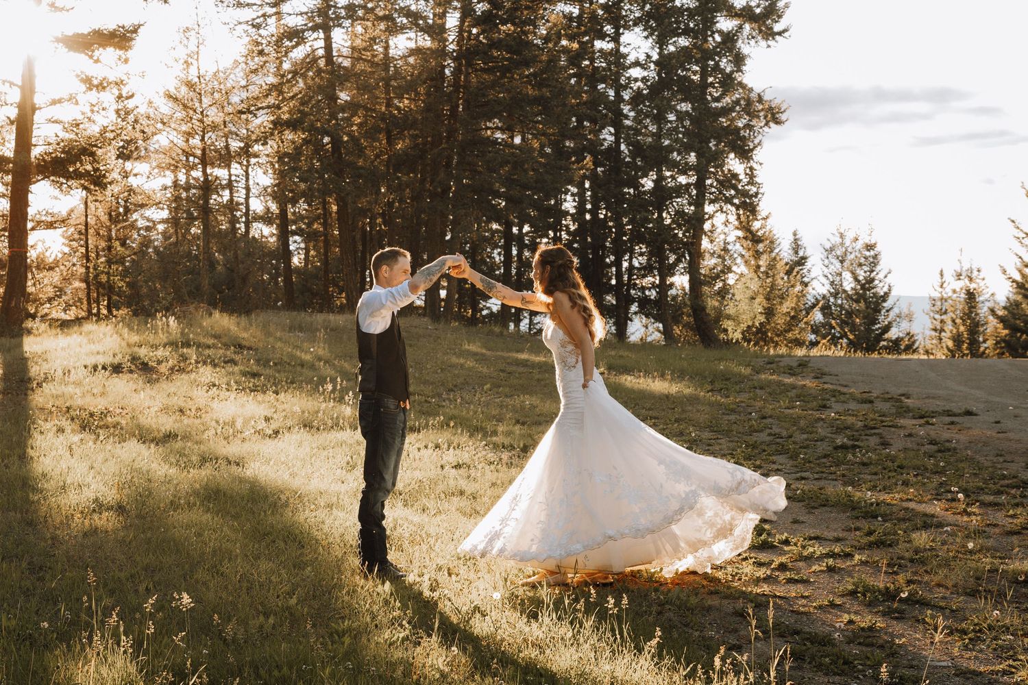 Bride and groom dancing in a sunlit meadow surrounded by tall pine trees in a romantic outdoor wedding setting.