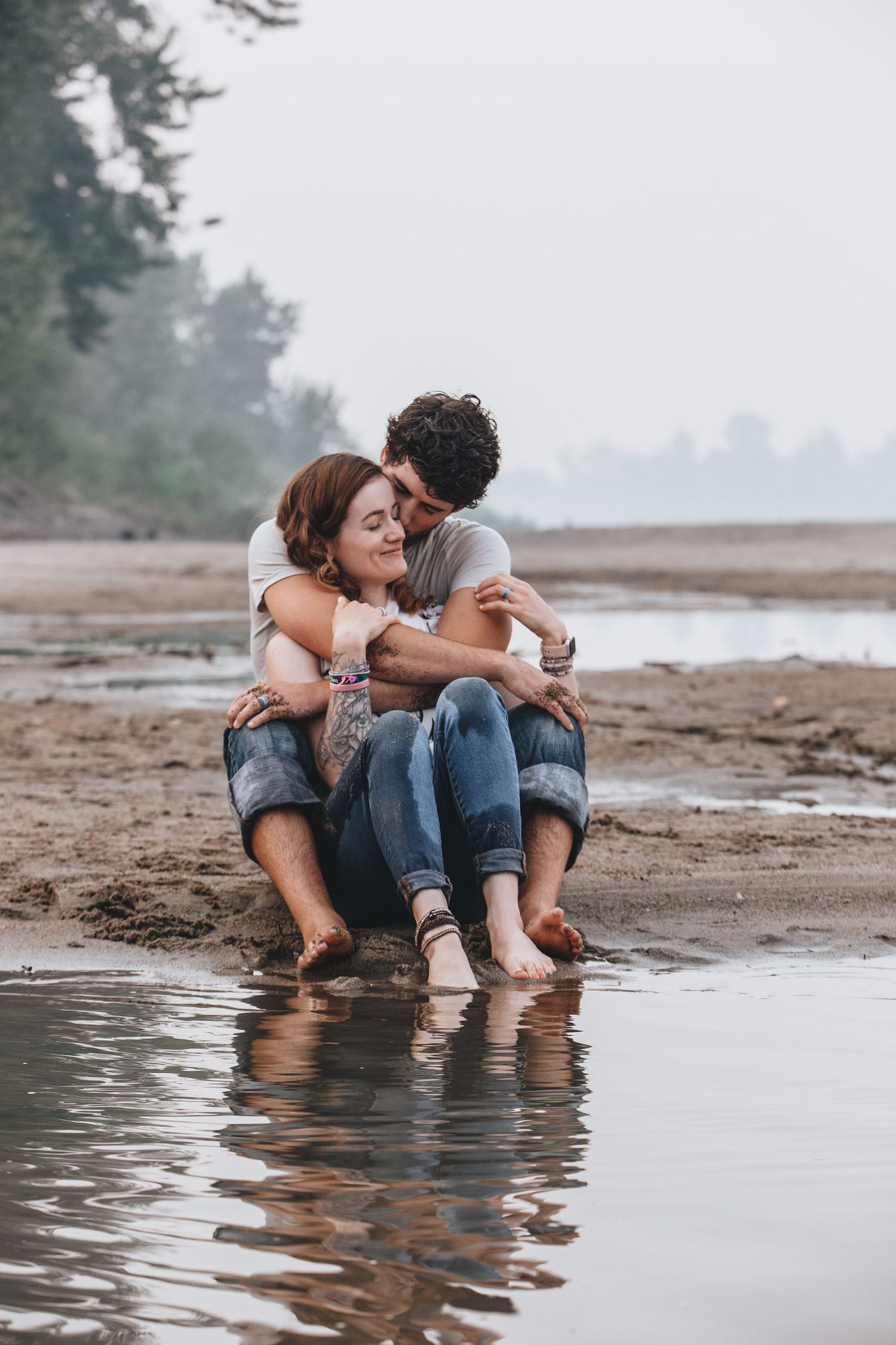 A couple sits at the edge of a misty beach shoreline with their feet in the water during sunset.