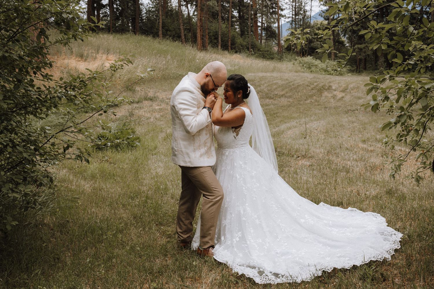 A couple in wedding attire shares an intimate moment in a rustic outdoor setting with tall grass and trees.