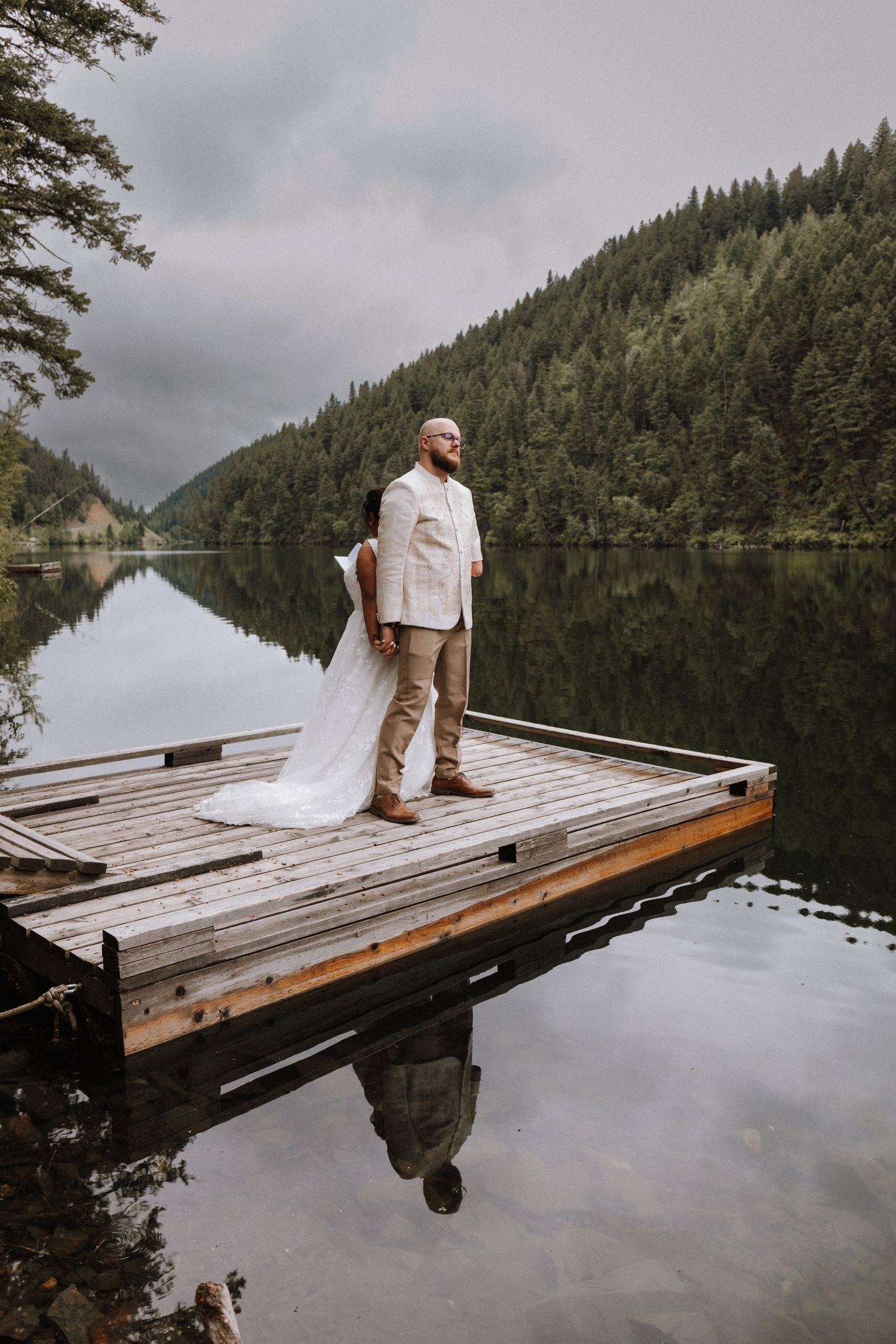 Couple does a first touch/ private vow on wooden dock overlooking serene mountain lake on cloudy day in Pacific Northwest.