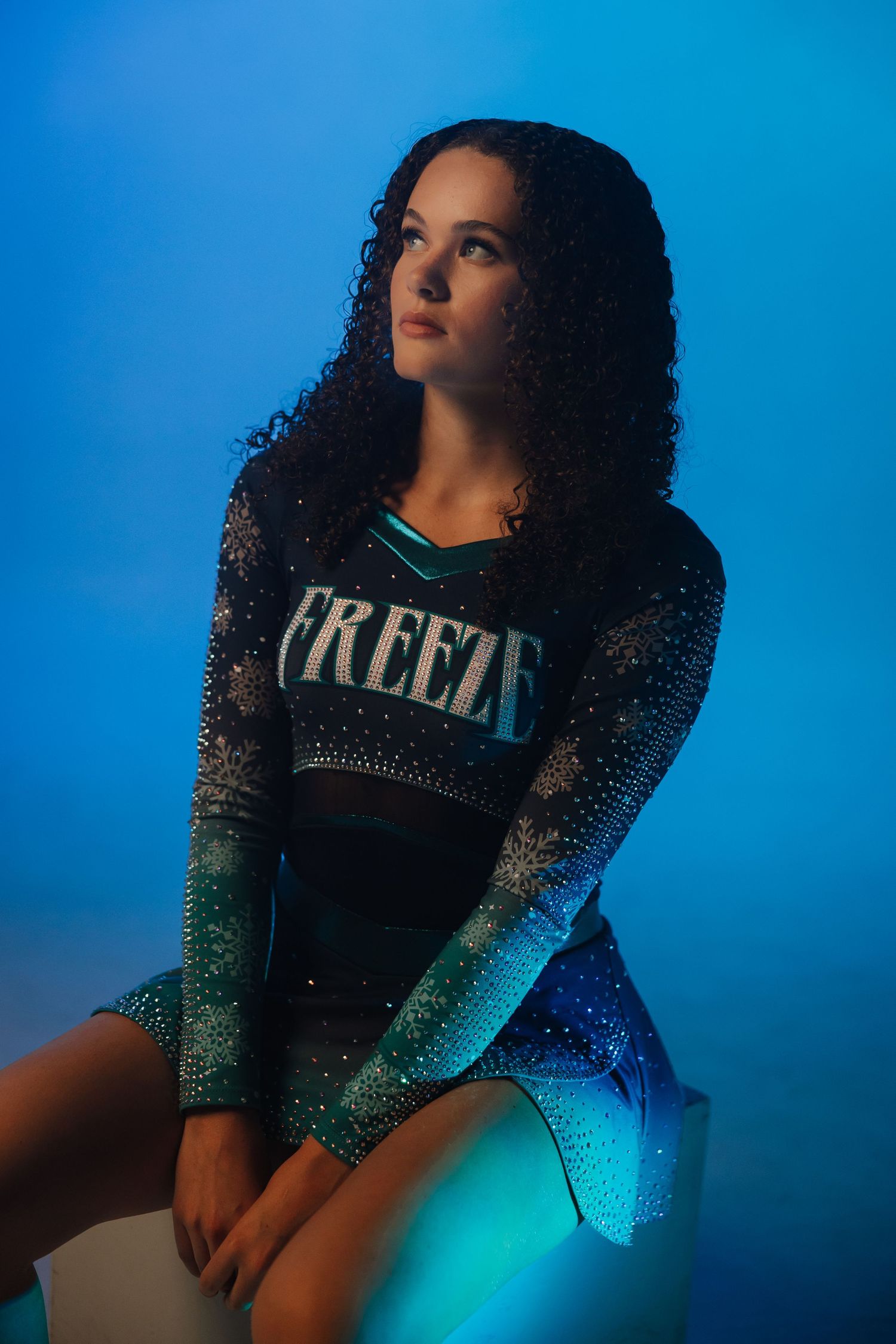 Young woman in a sparkly black and teal Freeze cheerleading uniform sits posed against a blue background.