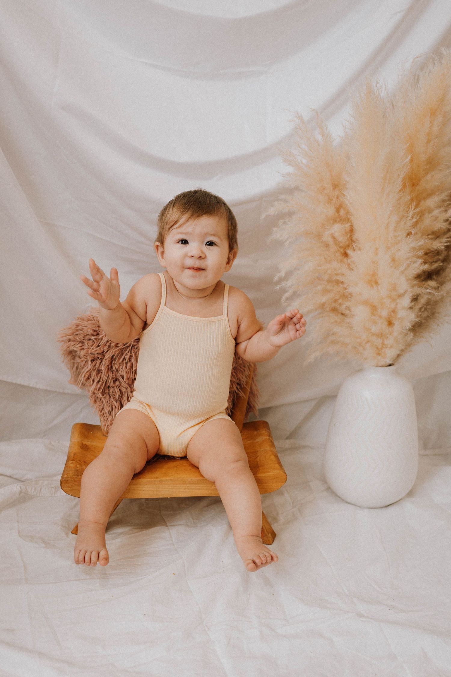 Baby in beige romper sitting on wooden bench with pampas grass decor.