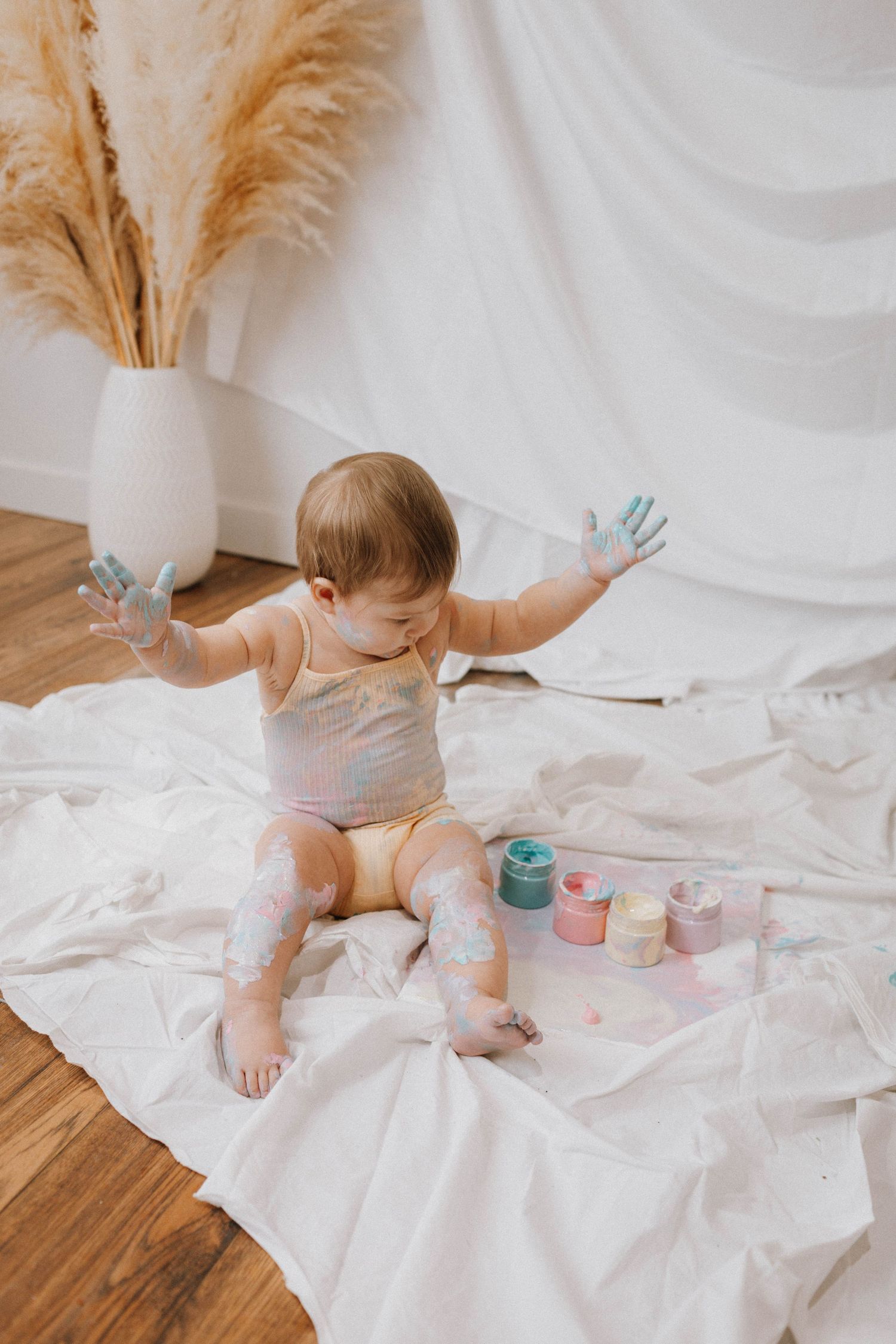 A baby sits on a white sheet wearing a gray onesie surrounded by paint jars and pampas grass decor.