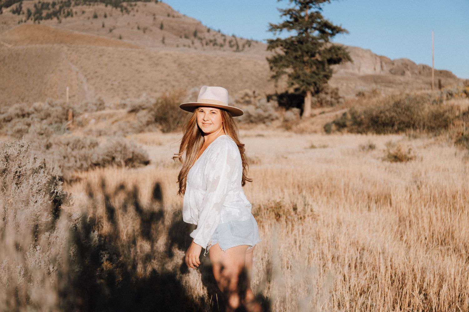 A person in a white dress and sun hat poses in a golden field with mountains in the background.