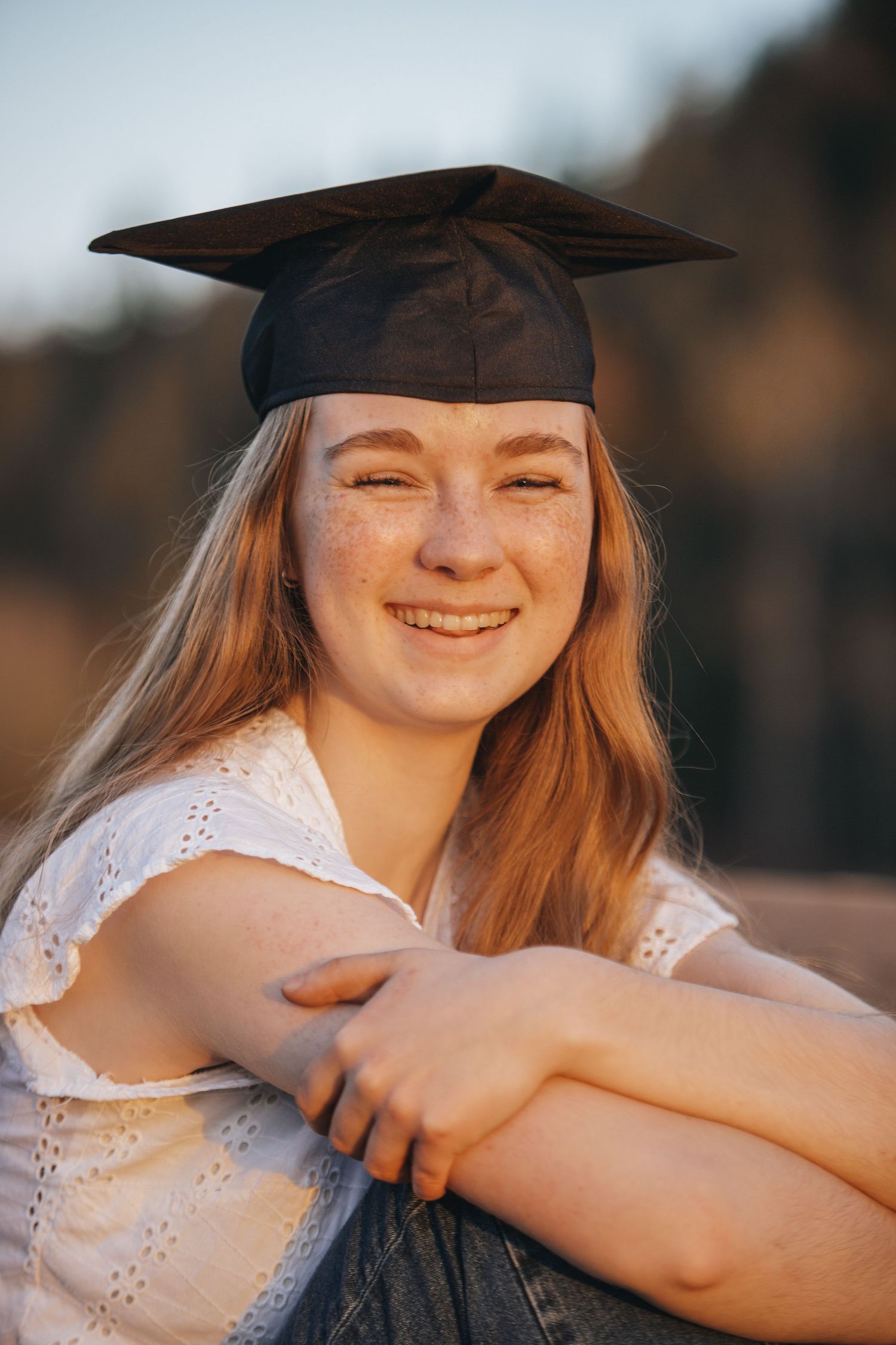 Graduate in white eyelet top and graduation cap poses outdoors at sunset.