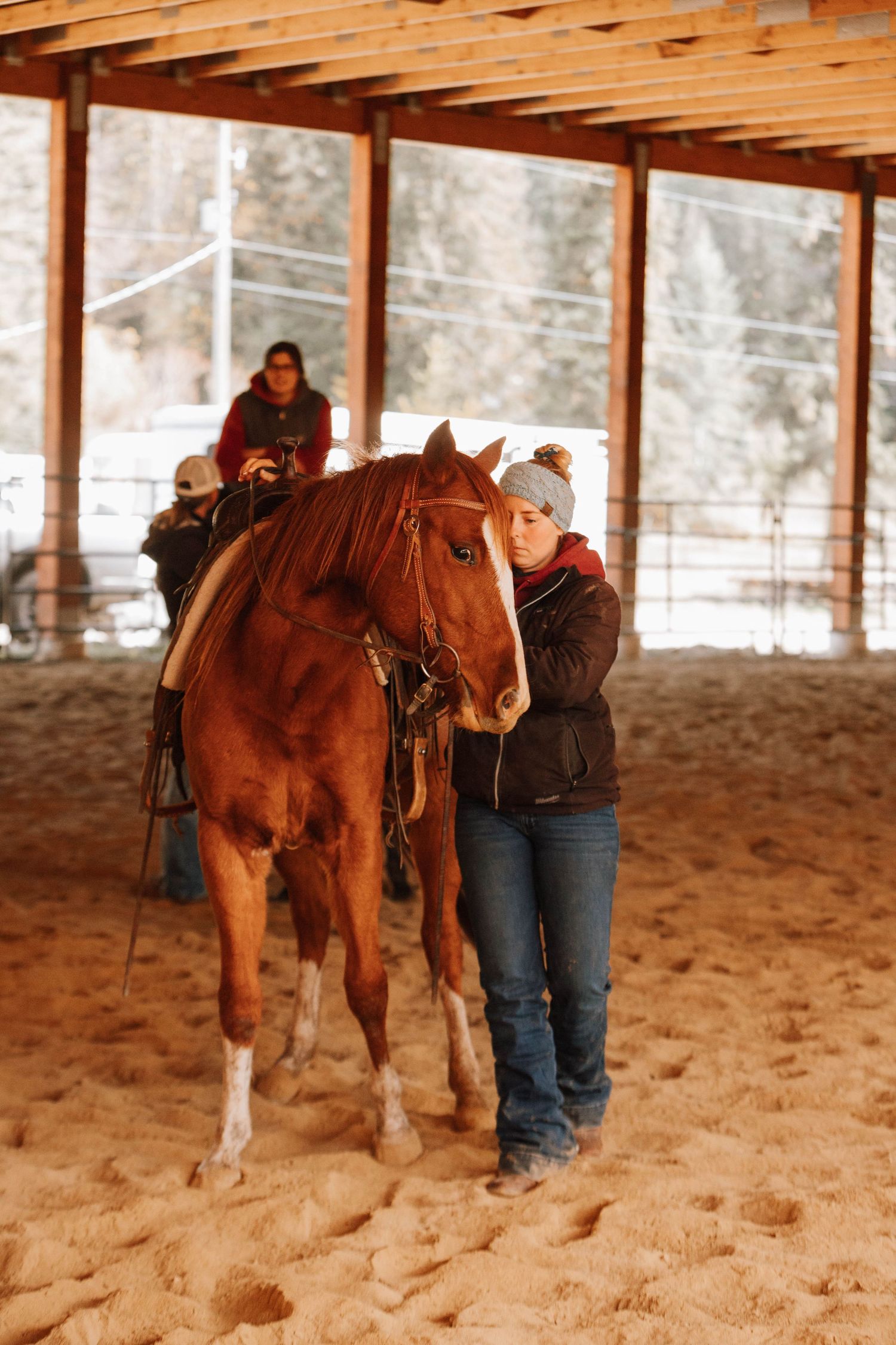 A person stands beside a saddled brown horse in a covered riding arena with sandy ground.