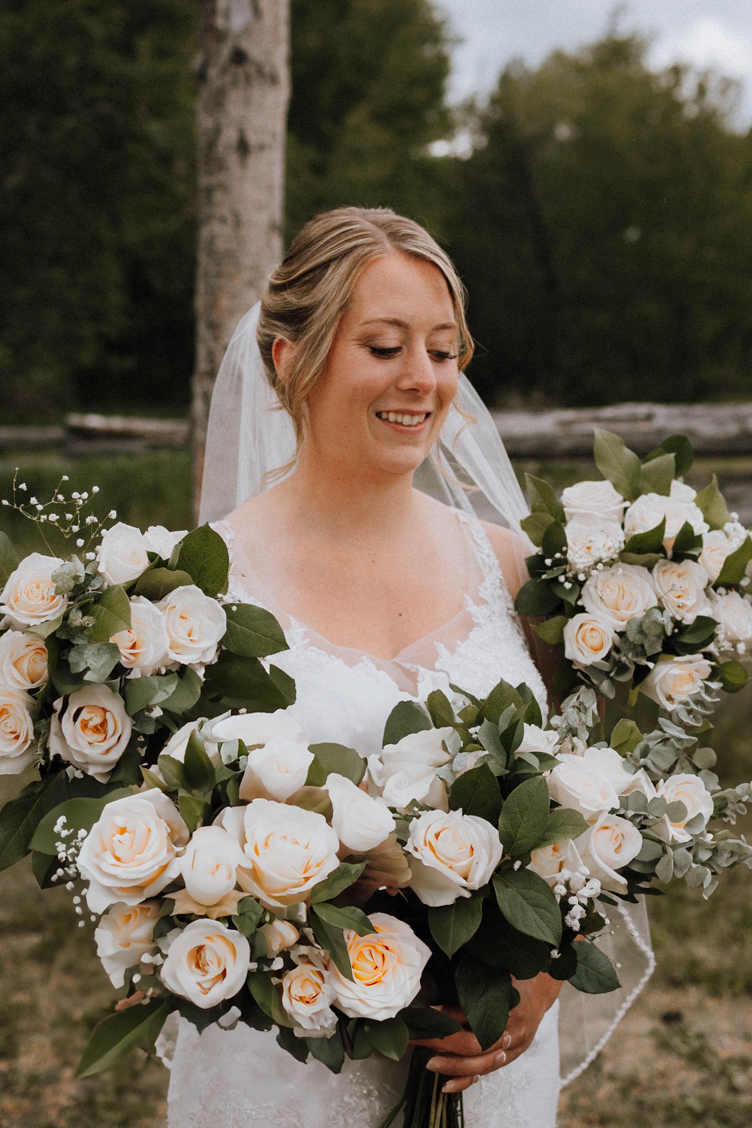Bride in white lace dress holds beautiful bouquet of cream roses and greenery at outdoor wedding.