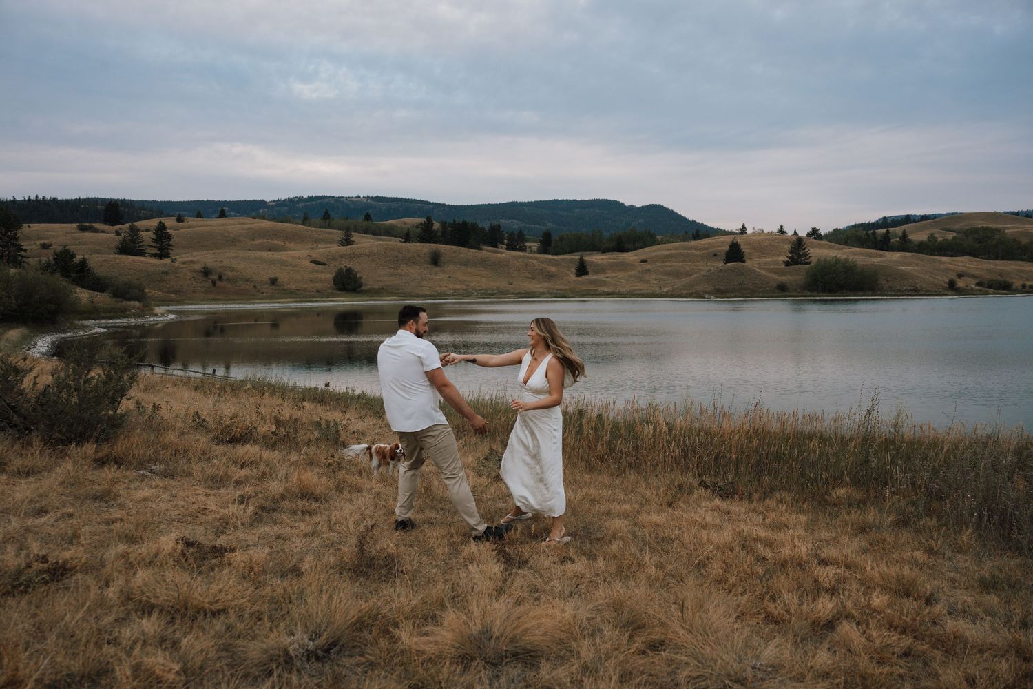 Couple dancing by a serene lake in golden fields with rolling hills at dusk.