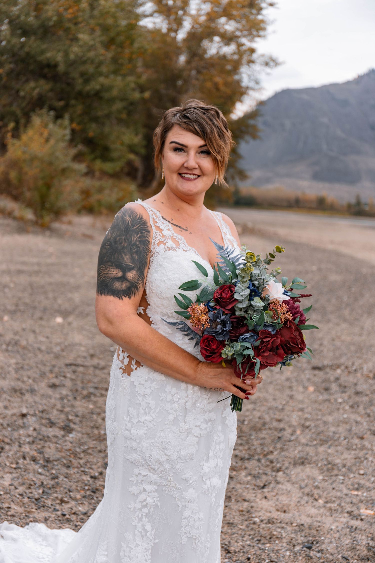 A bride in a lace gown poses with her burgundy and blue bouquet against an autumn mountain landscape.