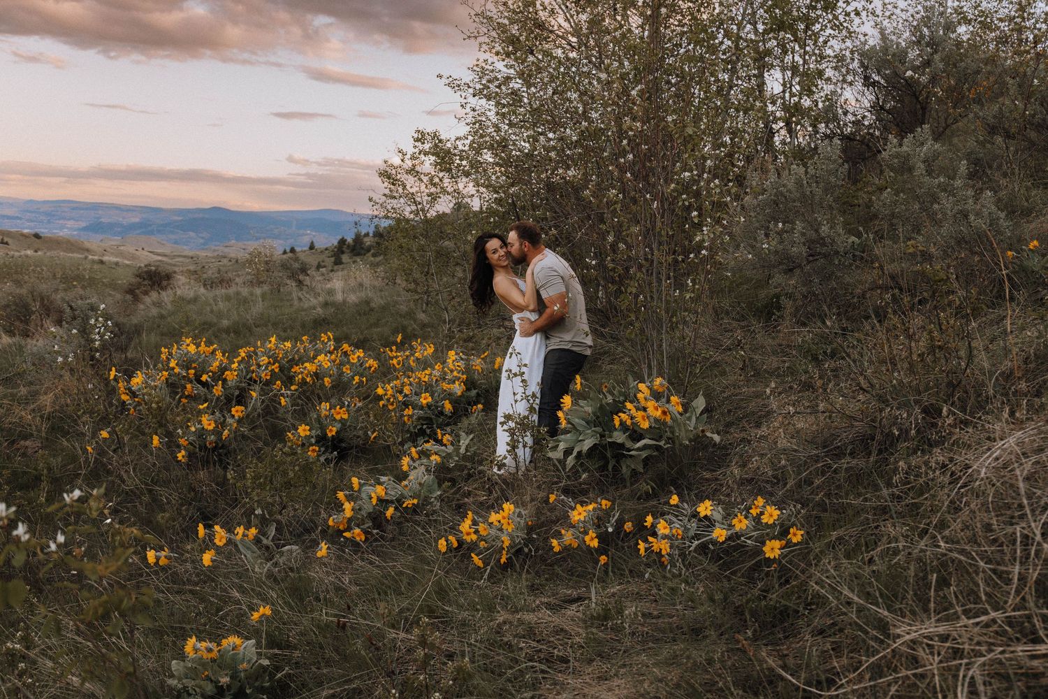 A couple embraces on a hillside at sunset surrounded by yellow wildflowers and mountain views in the distance.